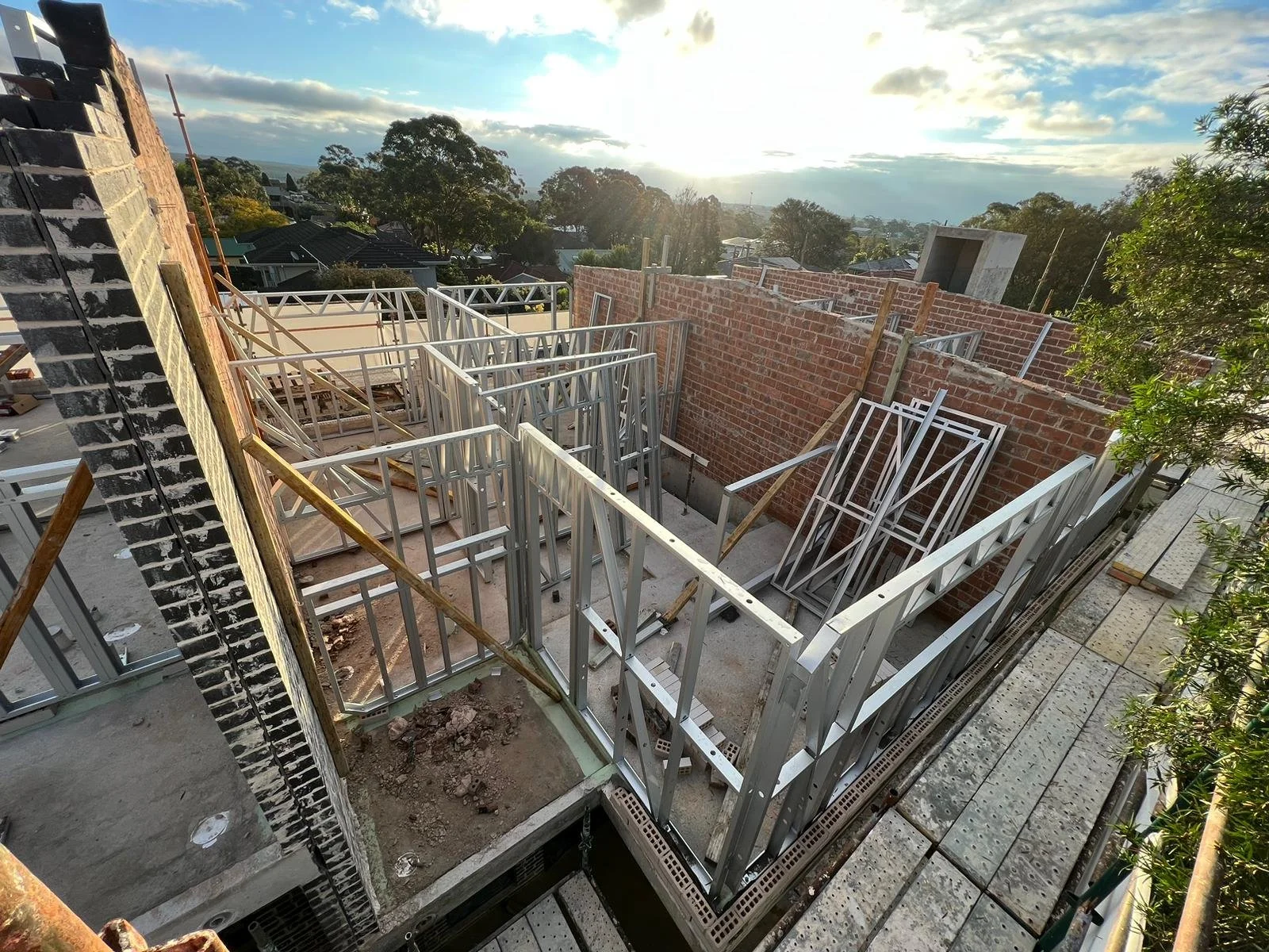 Construction site with metal framing for walls on an elevated platform, with a brick wall in the background and a scenic view of trees and sky at sunset.