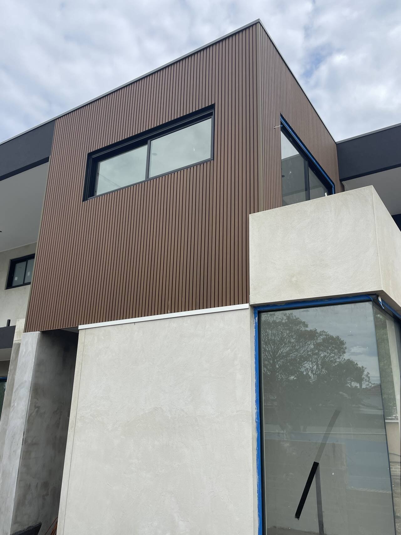 Modern multi-story building with brown vertical siding and large windows, featuring a white stucco wall and glass corner window, under a cloudy sky.