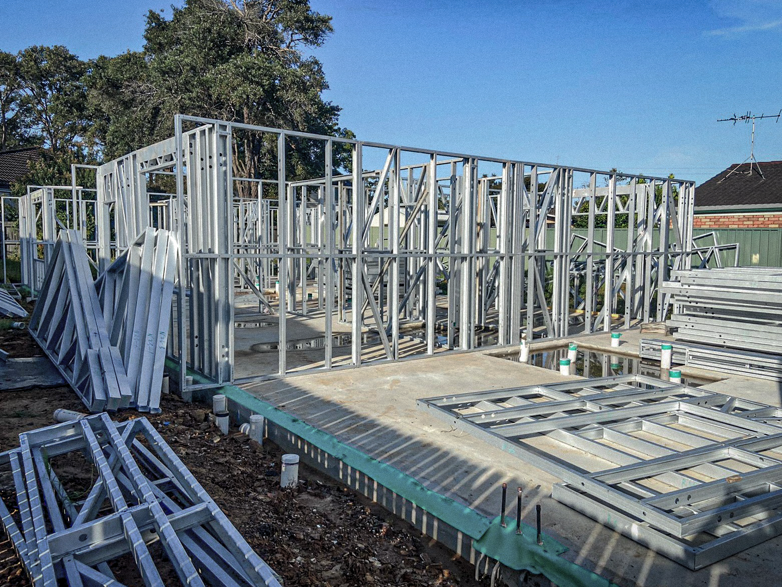 Under construction house framing with metal studs on concrete foundation, surrounded by building materials, under clear blue sky.