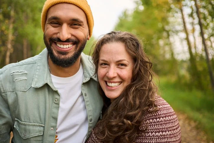 A happy young couple is shown in the woods while smiling and hugging. She has long brown hair and is wearing a sweater, he has on a light green shirt over a white t-shirt and is wearing a beanie.