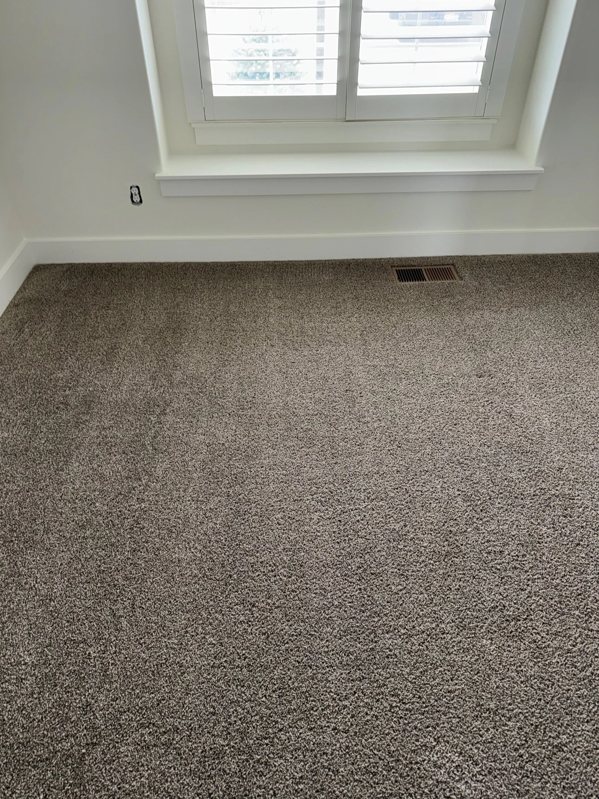 Empty room with beige carpet, a white wall with an electrical outlet, a window with white shutters, and a floor vent.