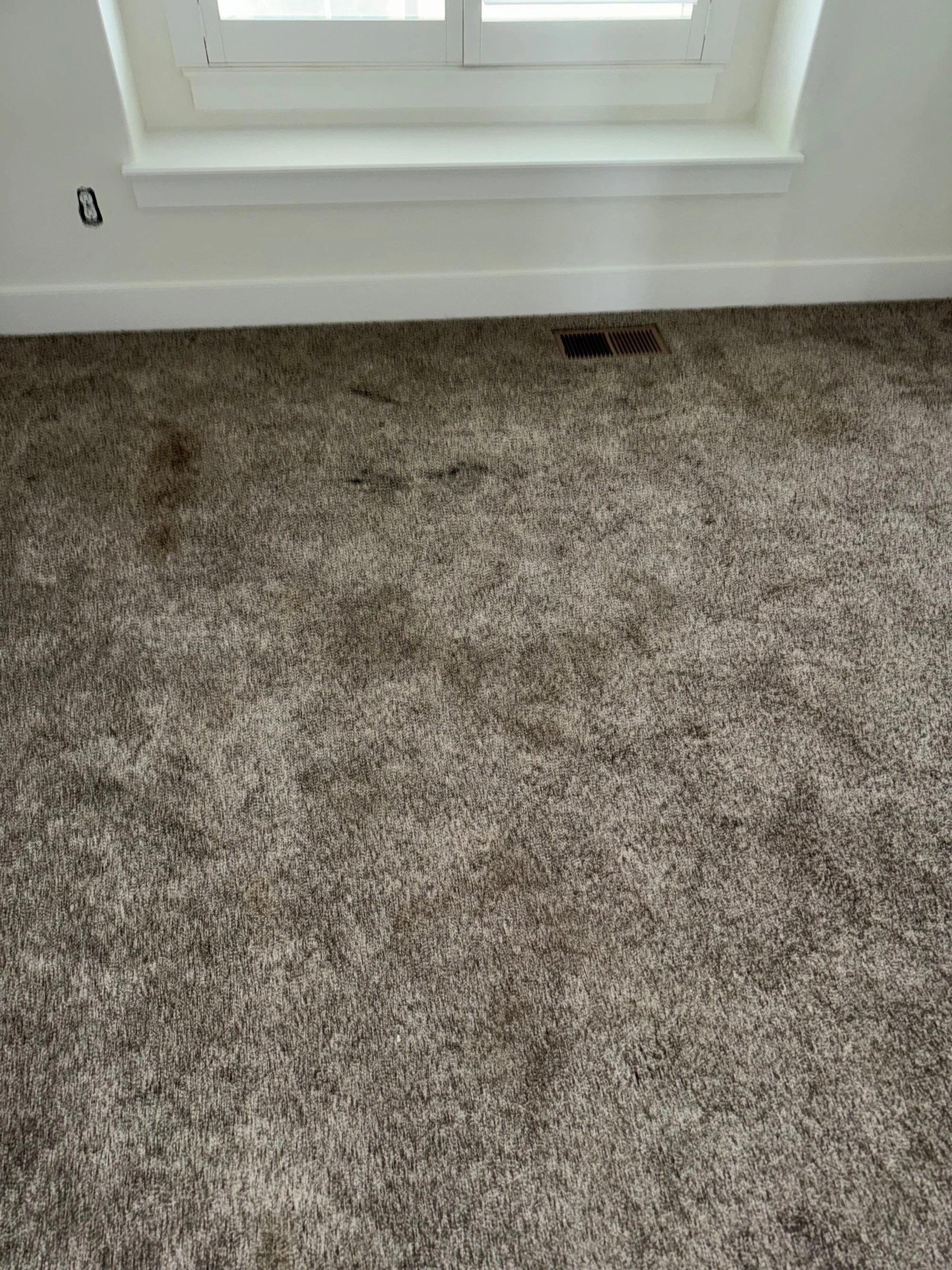 A beige carpeted floor with a darker stain near a white window sill. There is a heating vent near the window and an electrical outlet without a cover on the wall.