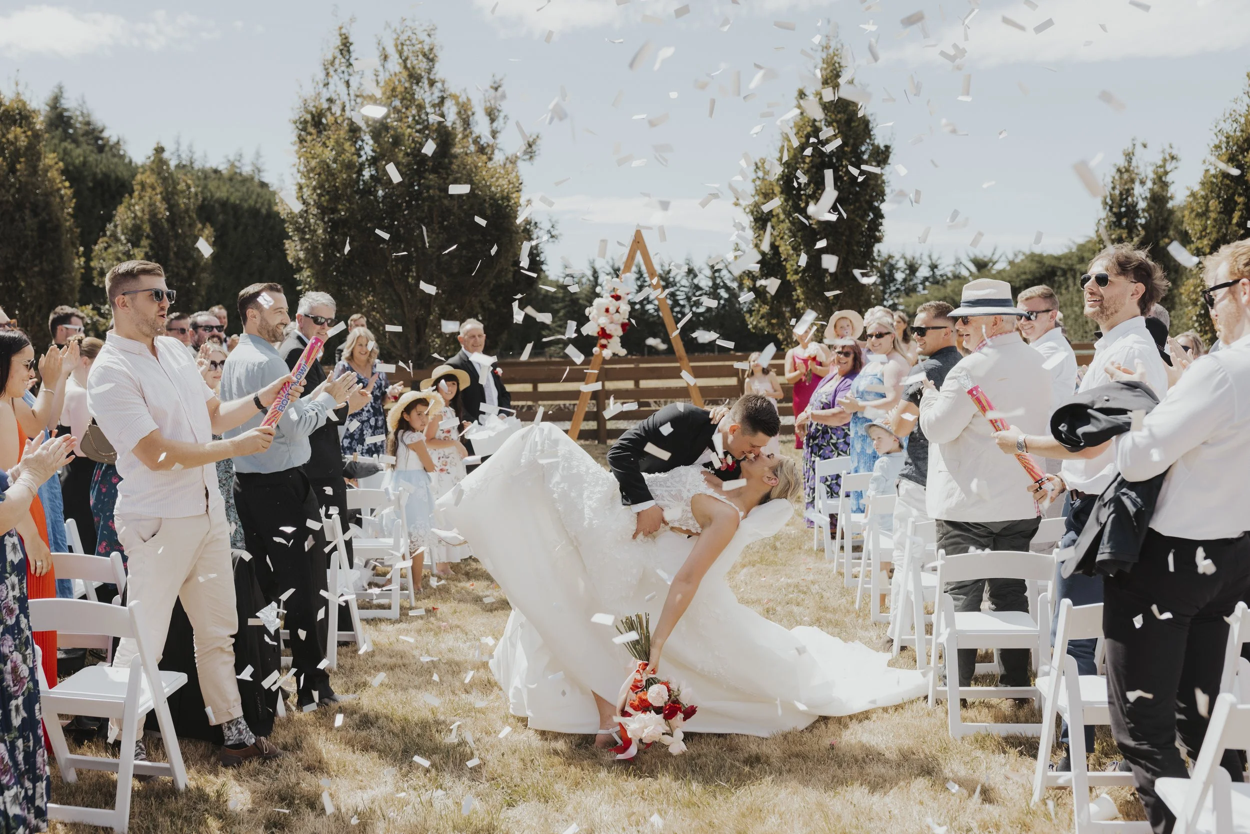 Bride and groom kissing with confetti as wedding guests cheers from Kylie and Nick's wedding at a golden field photographed by Sophie Isabella Photo