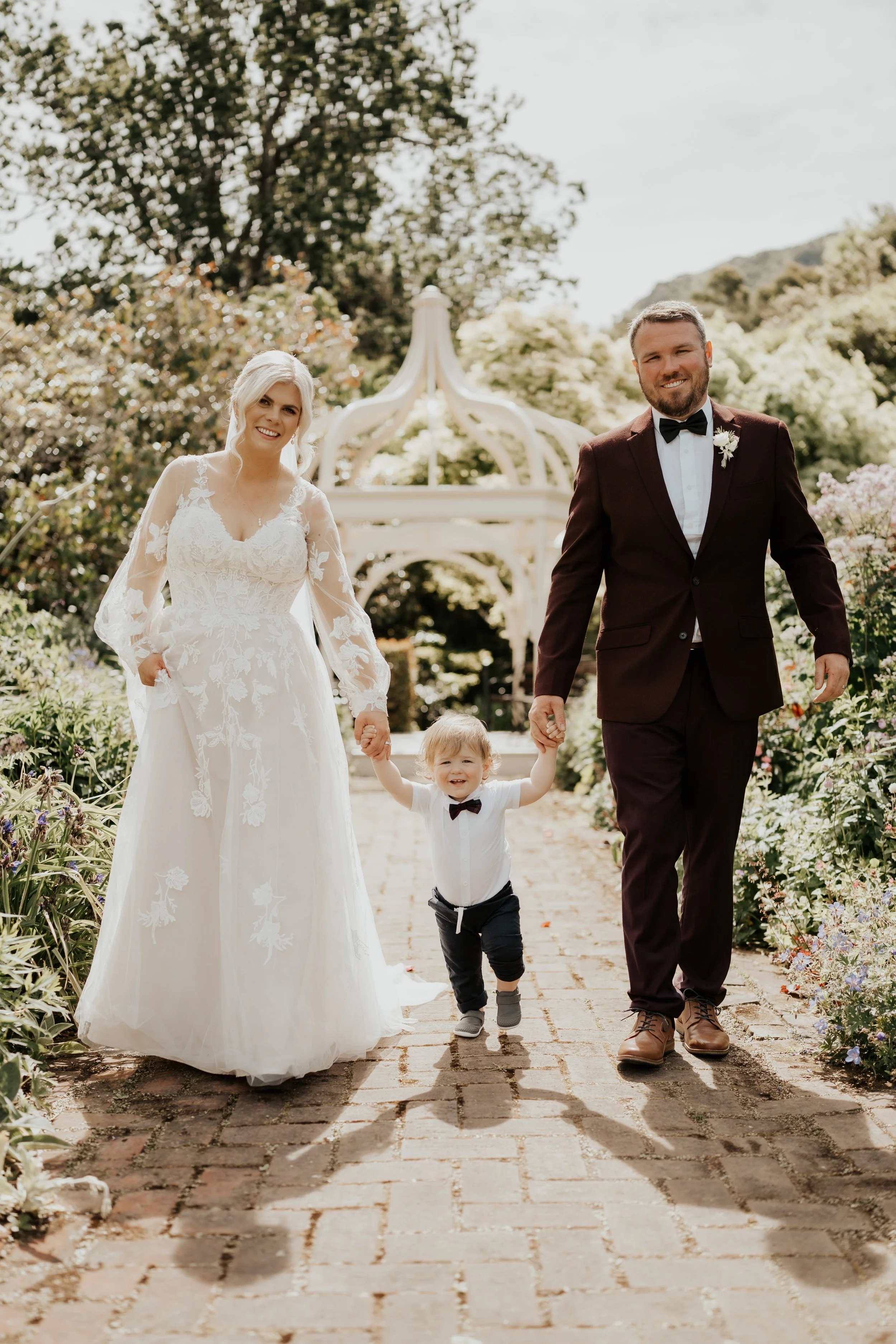Bride and groom with child walking away from white pergola in garden from Emma & Josh's wedding at Ōhinetahi Gardens and Milton St Substation photographed by Sarah Clements Photography