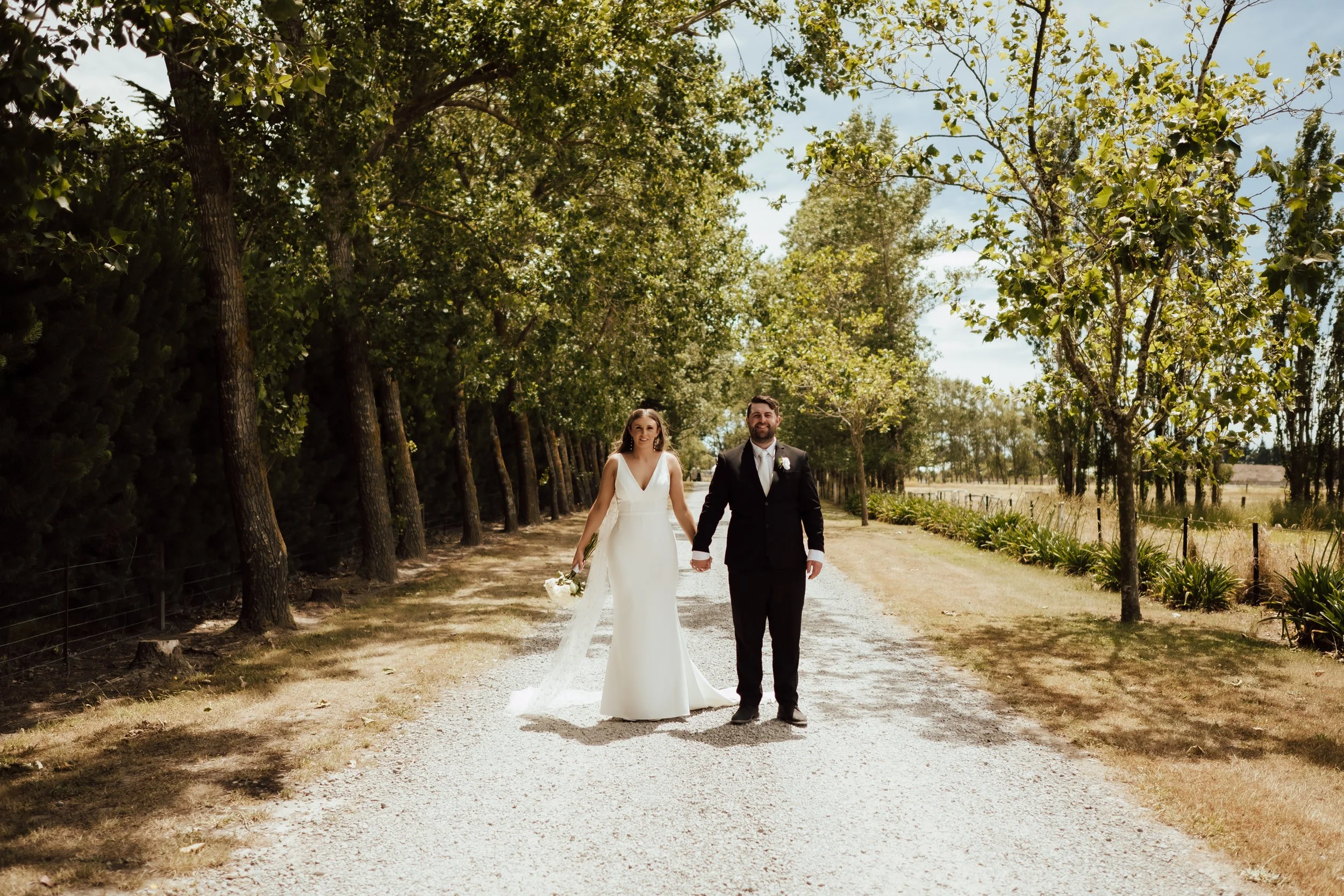 Bride and groom holding hands standing in tree lined drive from Lucy and Caleb's wedding at Larcomb Vineyard photographed by Tracey Allsopp
