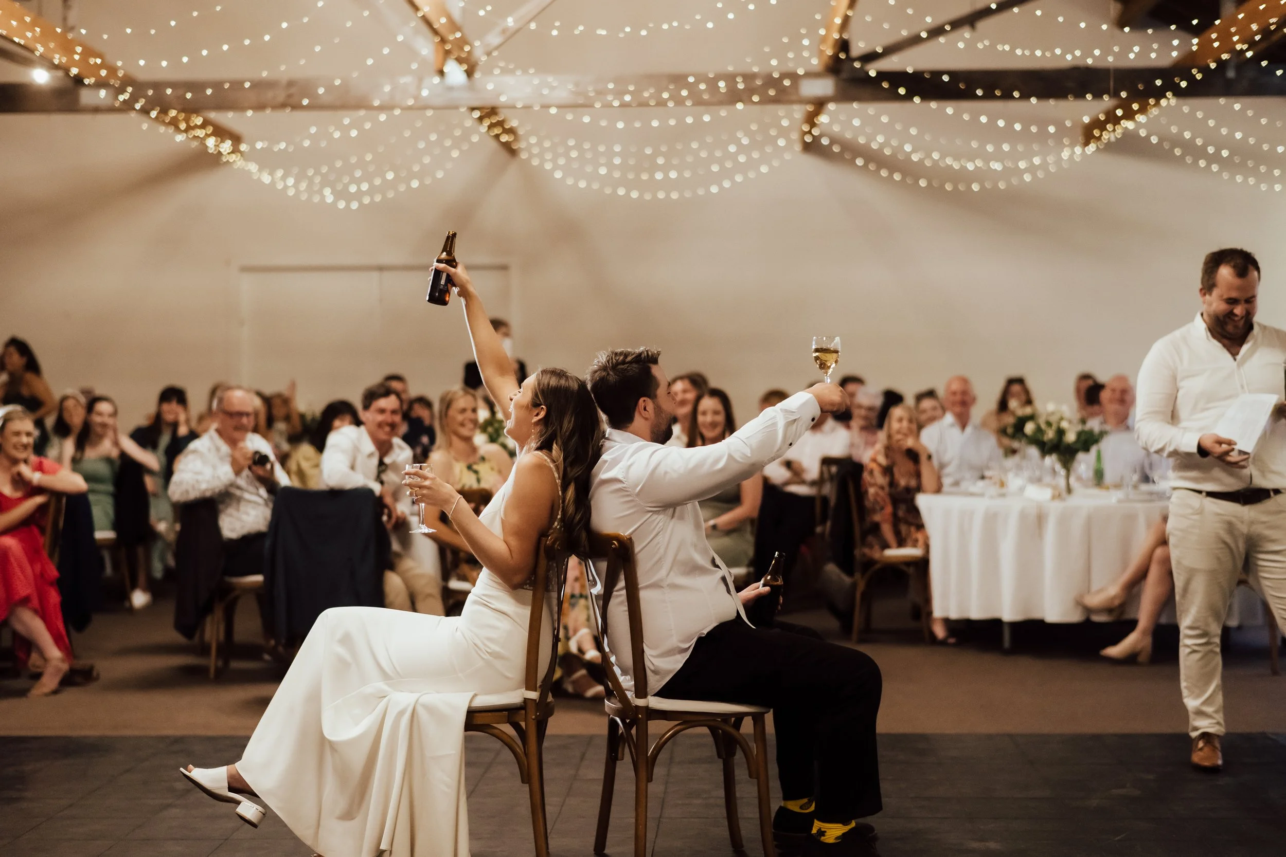 Bride and groom sitting in chairs with their backs to each other raising a toast while wedding guests watch from Lucy and Caleb's wedding at Larcomb Vineyard photographed by Tracey Allsopp