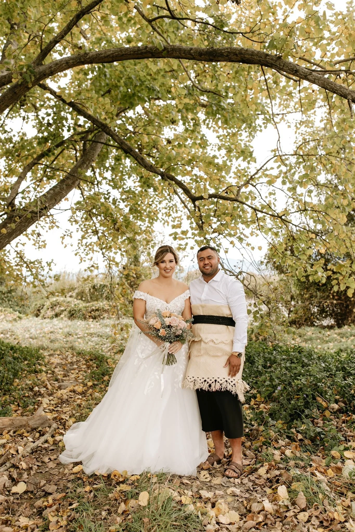 Bride wearing sweatheart neckline gown and groom wearing tupenu standing under autumnal tree from Hannah & Jarel's wedding at Seifried Estate photographed by Renee Edwards Photography