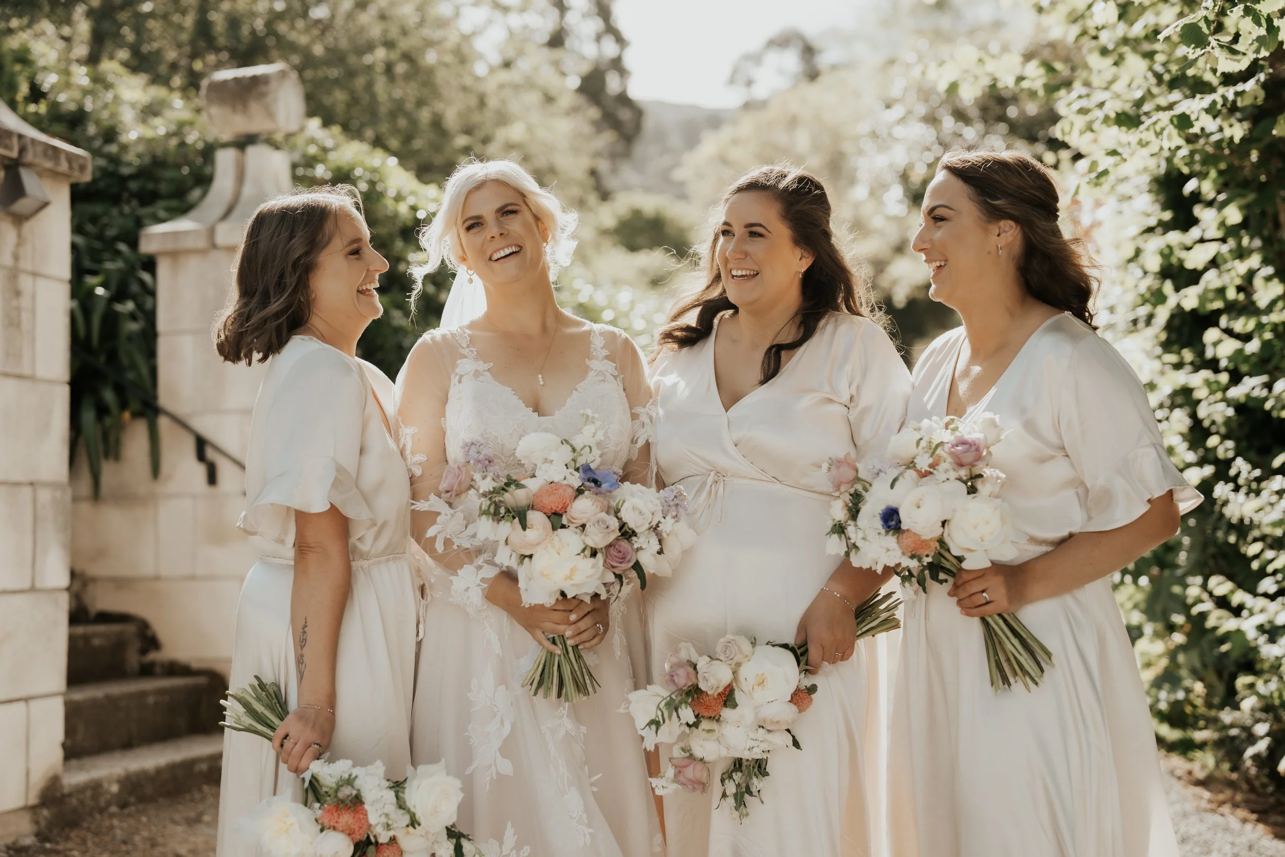 bridal party in front of stone gateway wearing white with white bouquets featuring pops of colour from Emma & Josh's wedding at Ōhinetahi Gardens and Milton St Substation photographed by Sarah Clements Photography