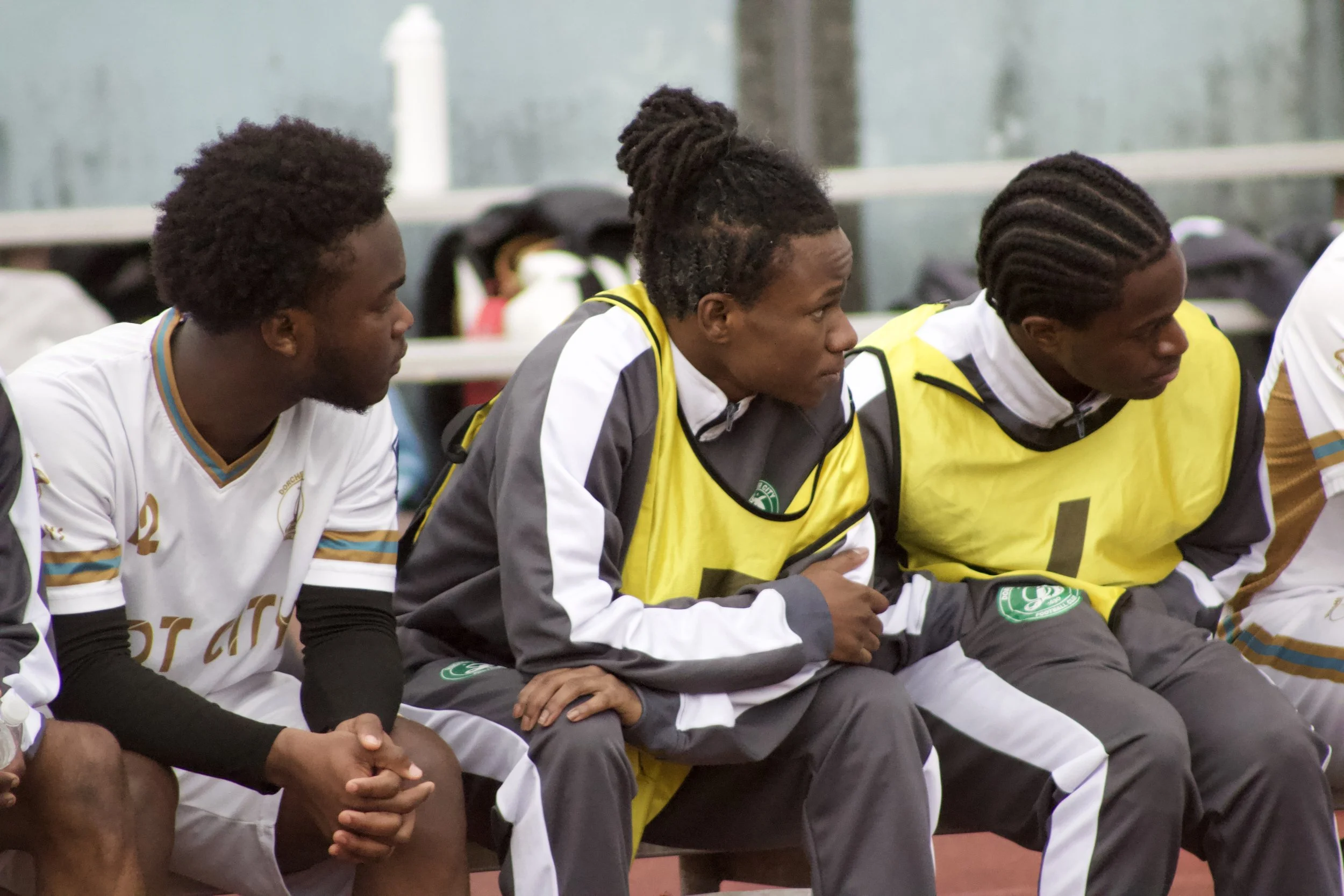 Three soccer players sitting on a bench during a game, one wearing a white jersey, two wearing yellow bibs, all with dreadlocked hair.