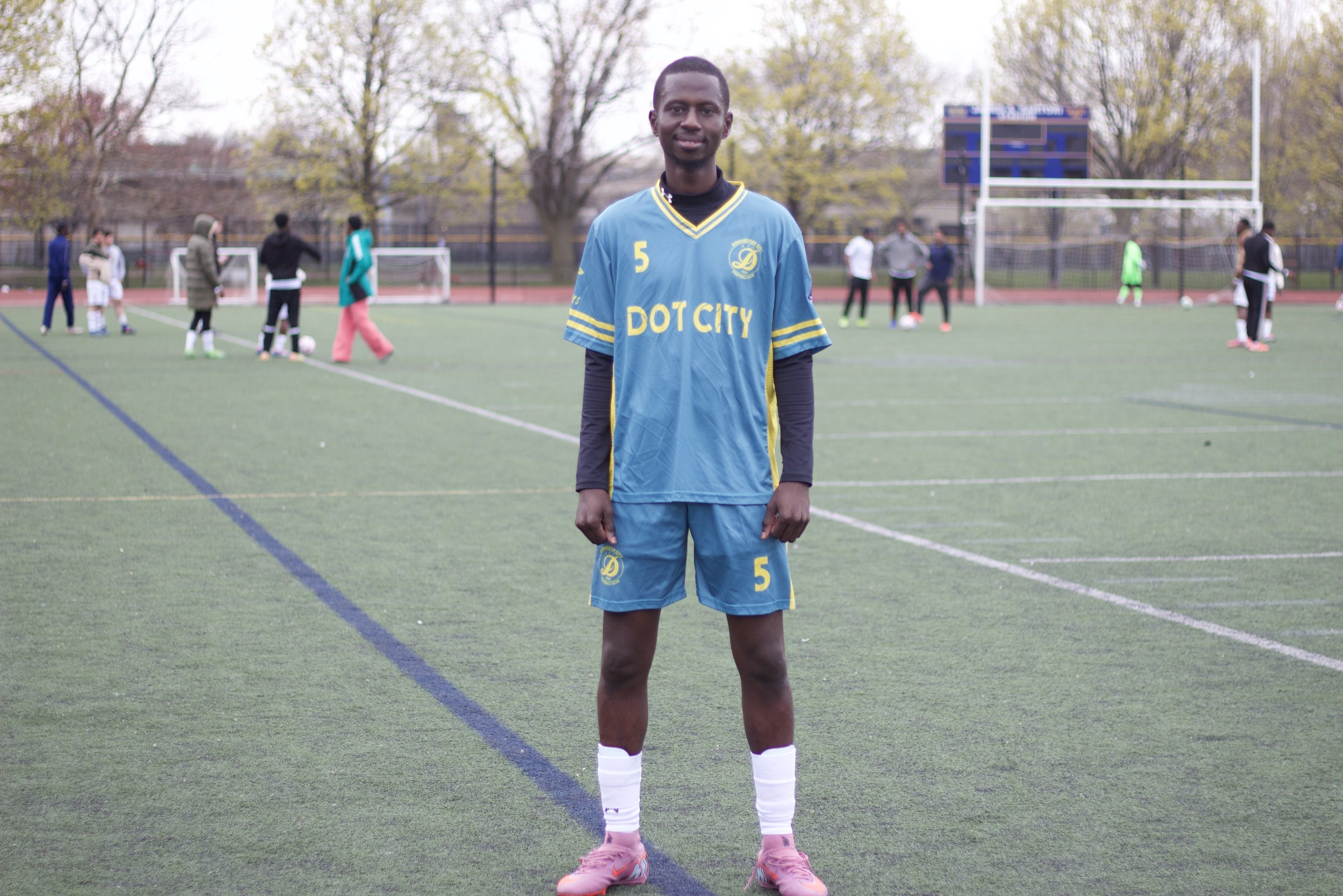 A young man standing on a soccer field wearing a blue sports uniform with the number 5, surrounded by other players practicing in the background, and trees in the distance.