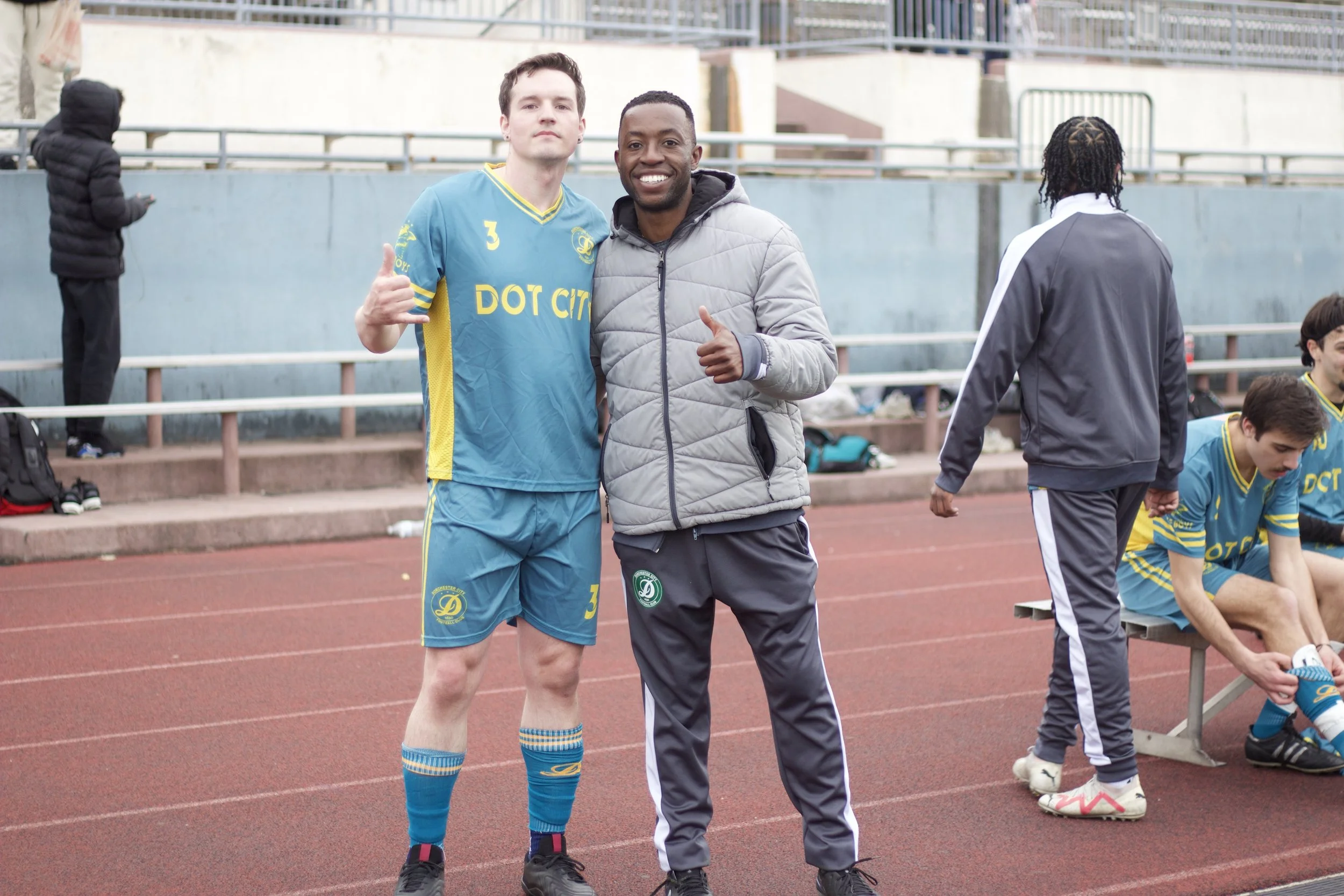 Two men standing on a track field, one in a blue sports uniform and the other in casual sportswear, smiling and giving a thumbs-up. Several others are in the background, some seated and some standing.