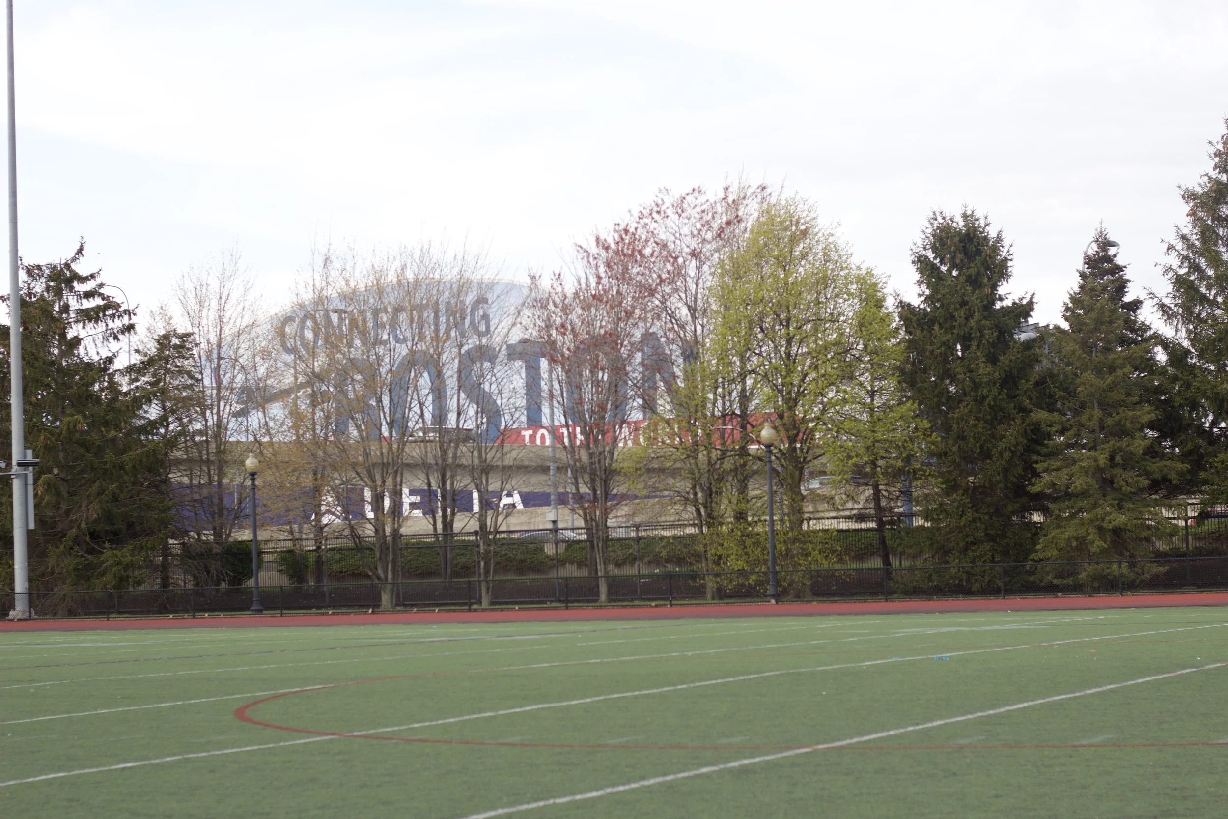 An outdoor sports field with a red and green track and a green playing surface, against a backdrop of trees and hills under cloudy sky.