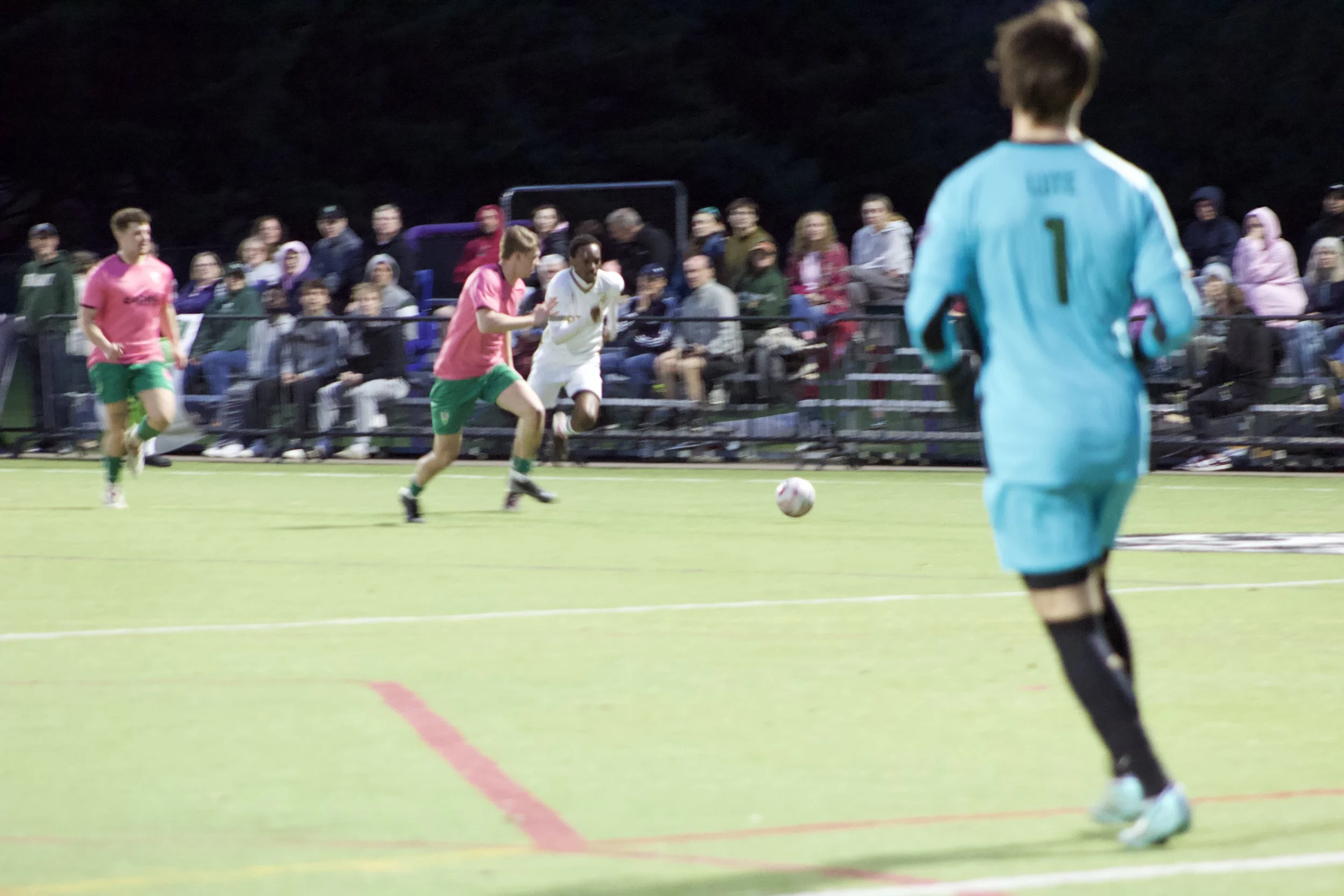 A soccer match is taking place on a field with players in pink and white uniforms, and a goalkeeper in light blue. Spectators are watching from the stands at night.