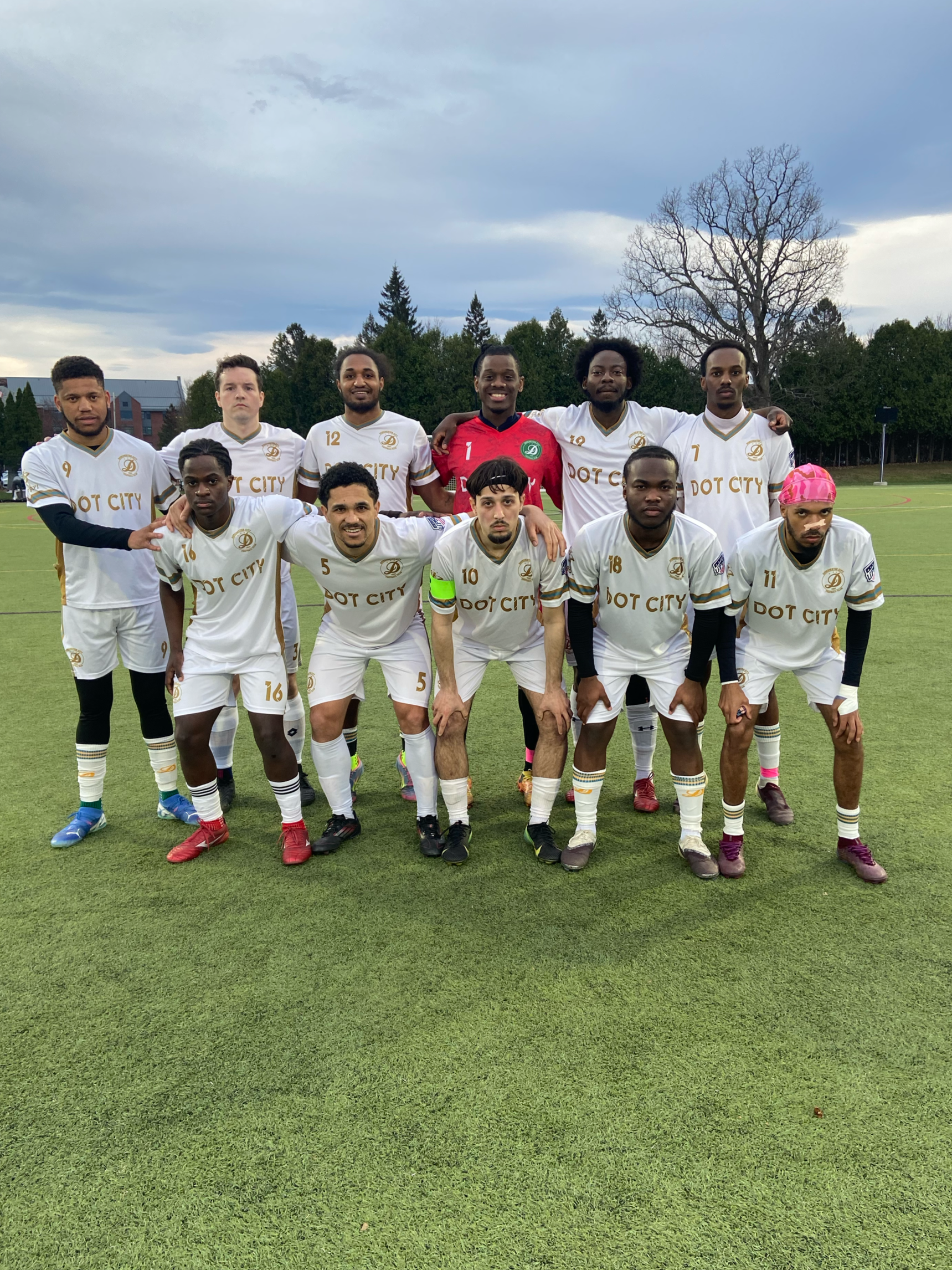 A group of eleven men in soccer uniforms stands on a soccer field for a team photo. They are wearing white jerseys with gold and black accents, and some are wearing black, red, or purple cleats. The central goalkeeper is wearing a red jersey. The pla