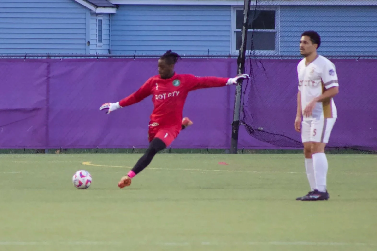 A soccer player in a red uniform kicking a ball while another player in a white uniform stands nearby on the field.