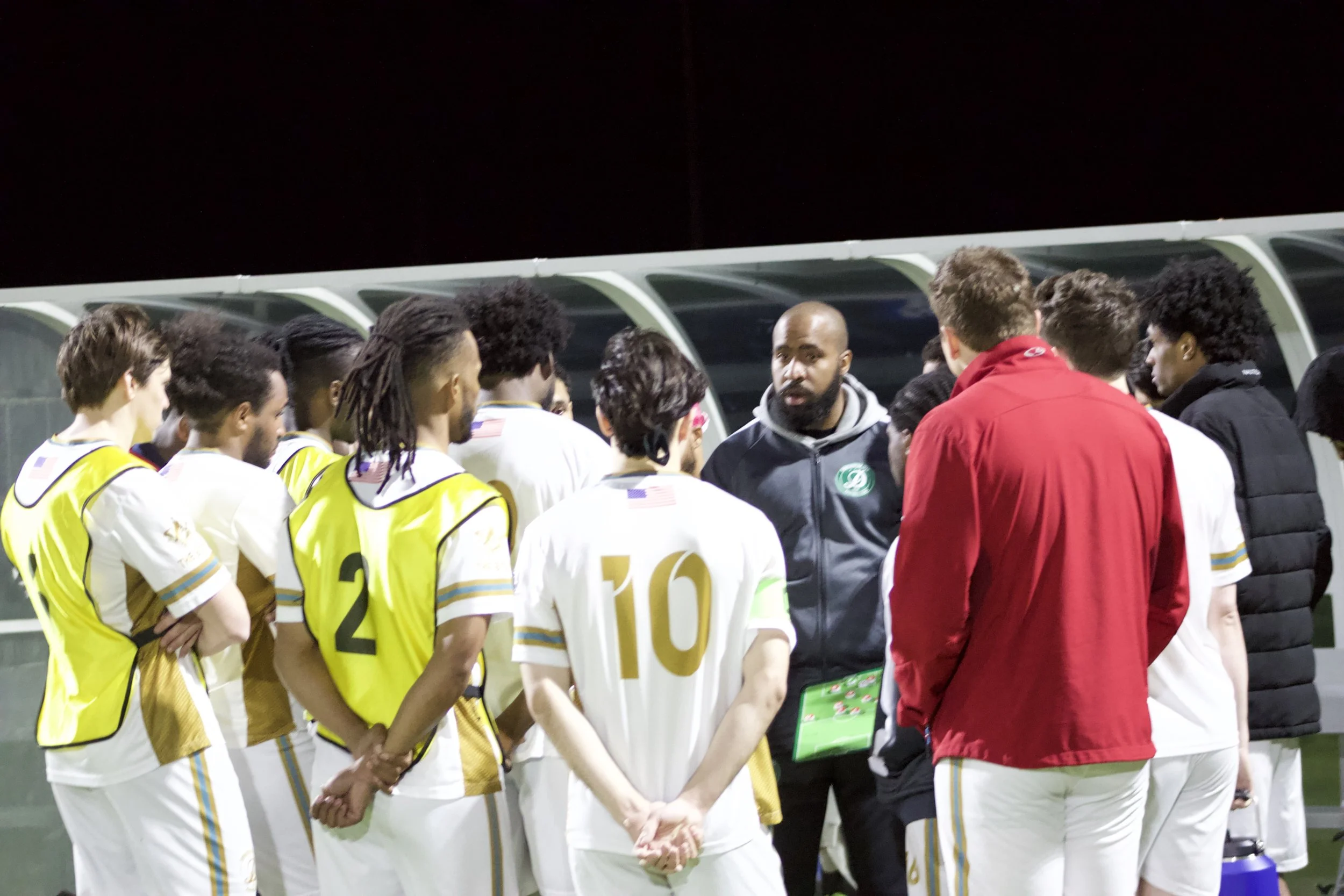 Soccer team members in white and yellow uniforms huddle around their coach, who is giving instructions on the sideline of a soccer field at night.