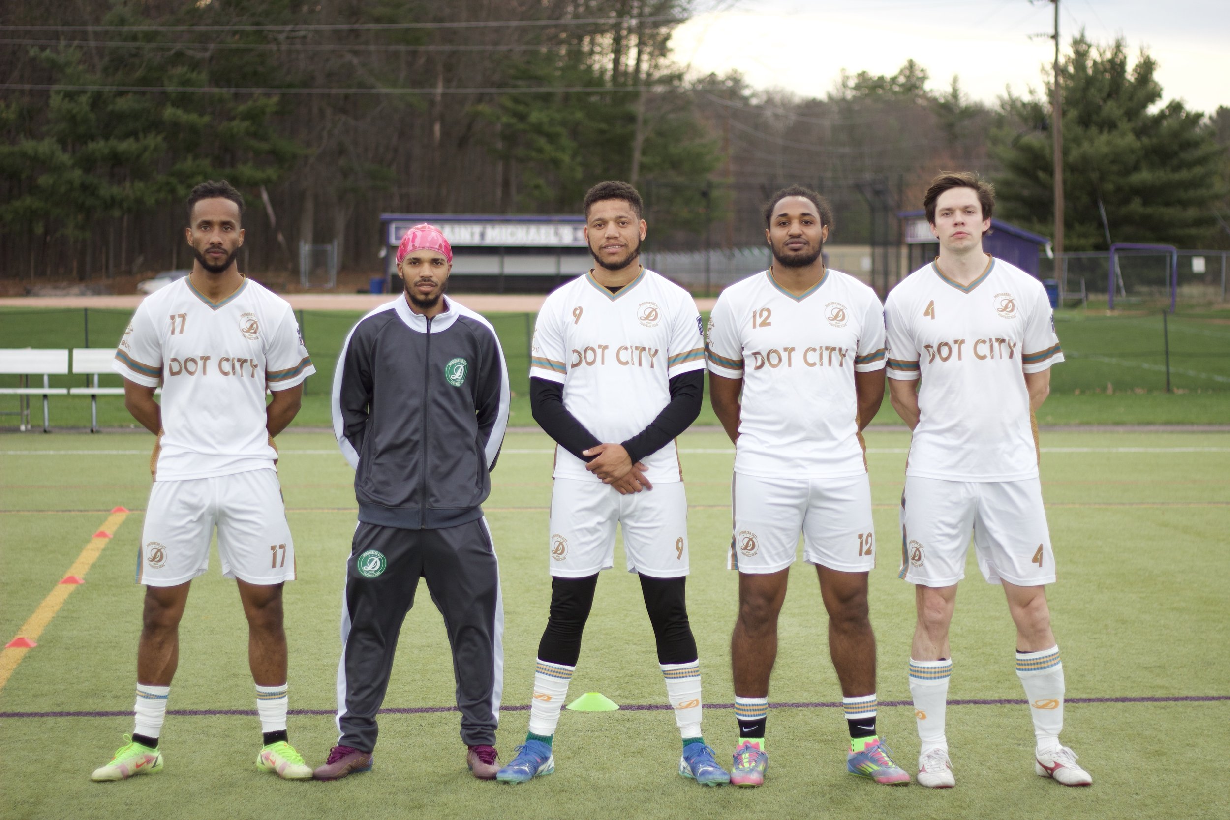 Five male soccer players standing side by side on a soccer field, wearing white jerseys with gold and blue accents, and a coach in a black jacket standing in the middle. They are outdoors, with trees and a signboard in the background.