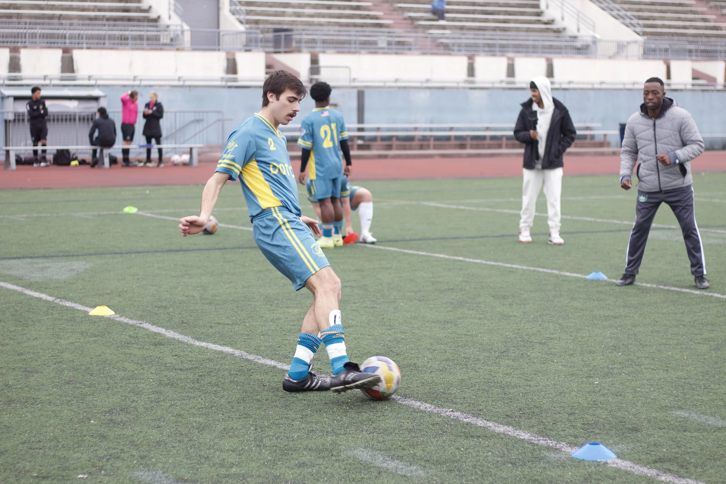 A young man wearing a blue and yellow soccer uniform practicing on a field with a soccer ball, with other people and players in the background.