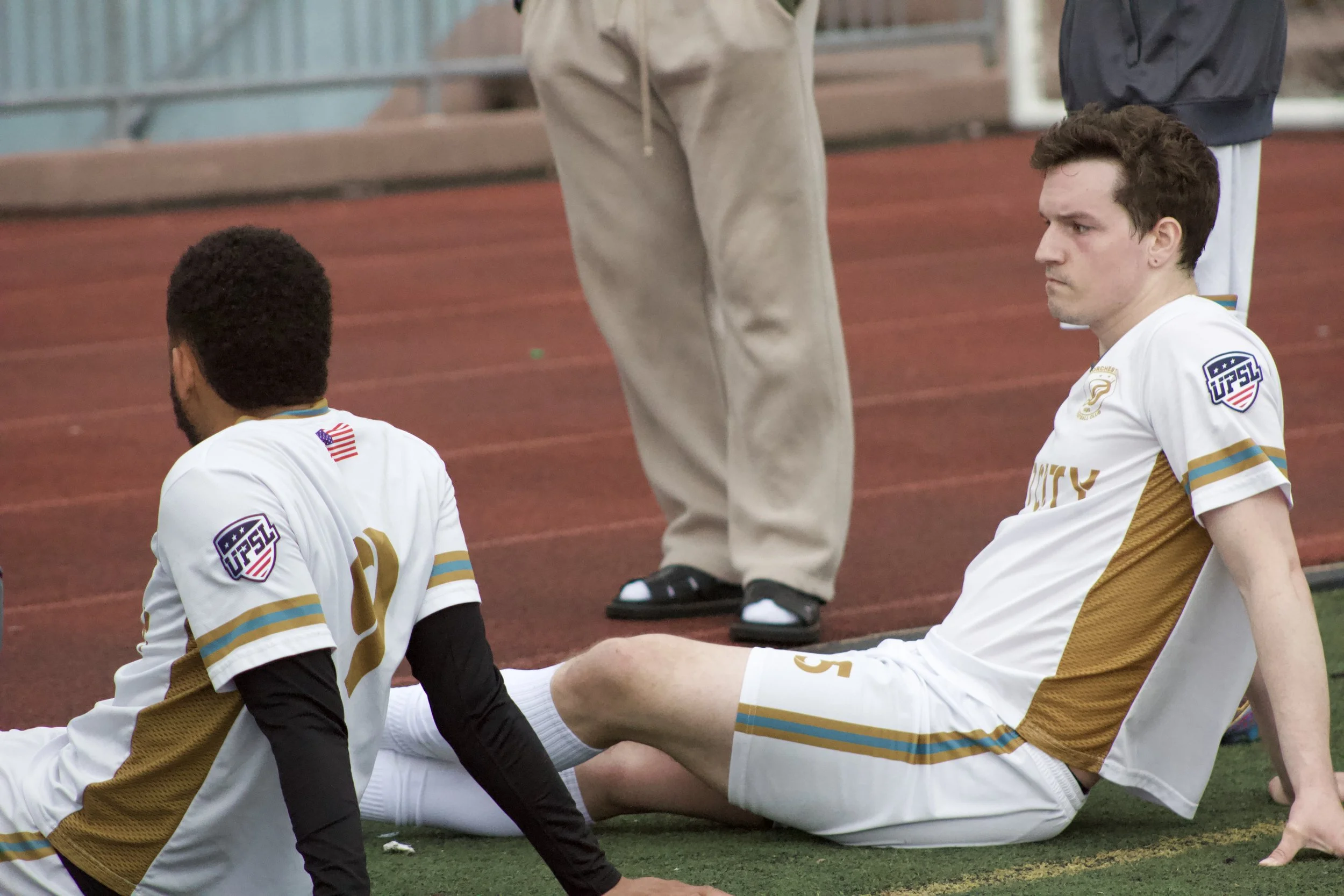 Two soccer players sitting on the field, wearing white and gold jerseys with patches, near a red running track and a fence. One is African American with short curly hair, and the other is Caucasian with short brown hair.