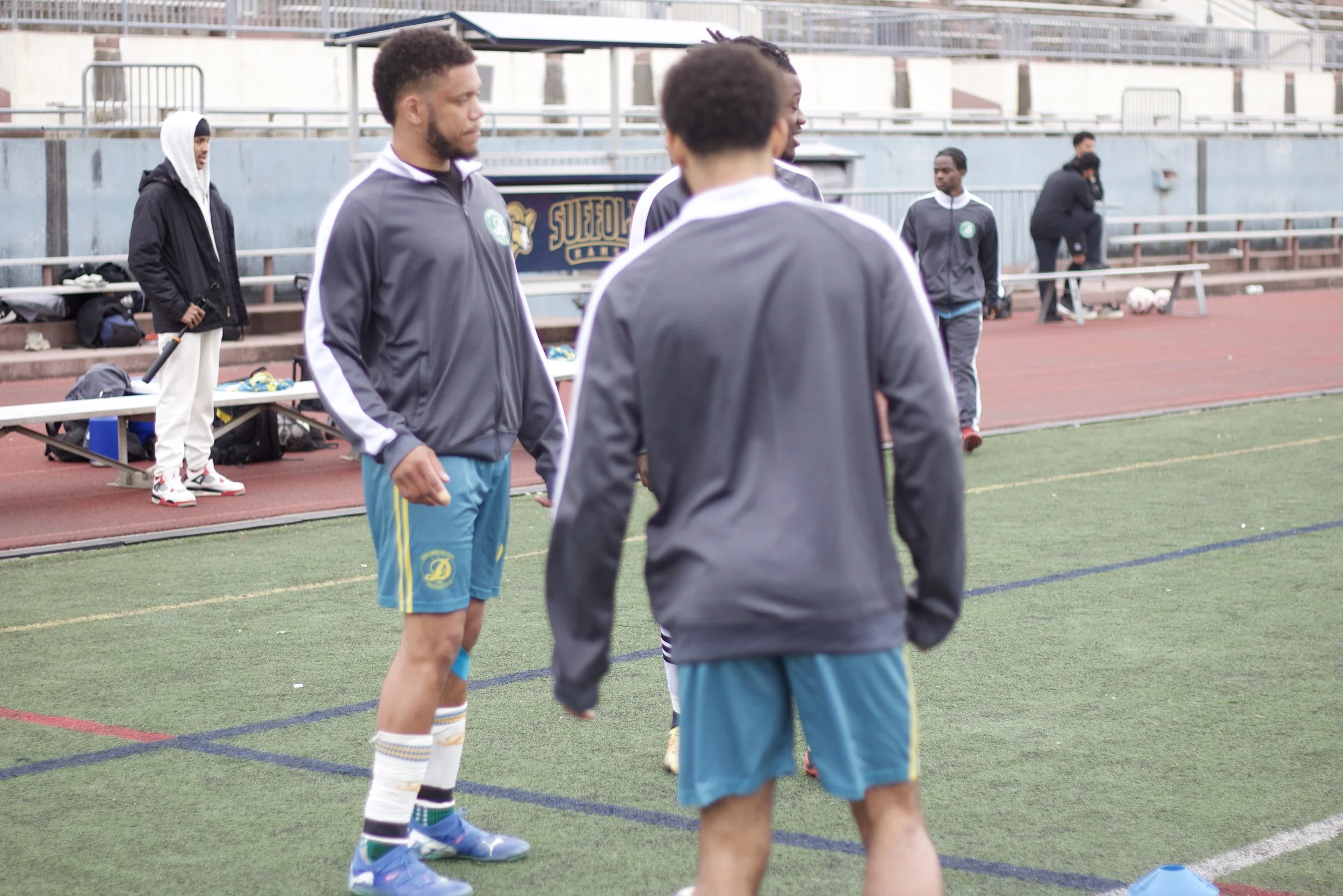 A group of young men in athletic clothing standing on a running track, preparing for a sports practice or game at an outdoor stadium. Some are wearing jackets and shorts, with a few wearing cleats and compression socks.