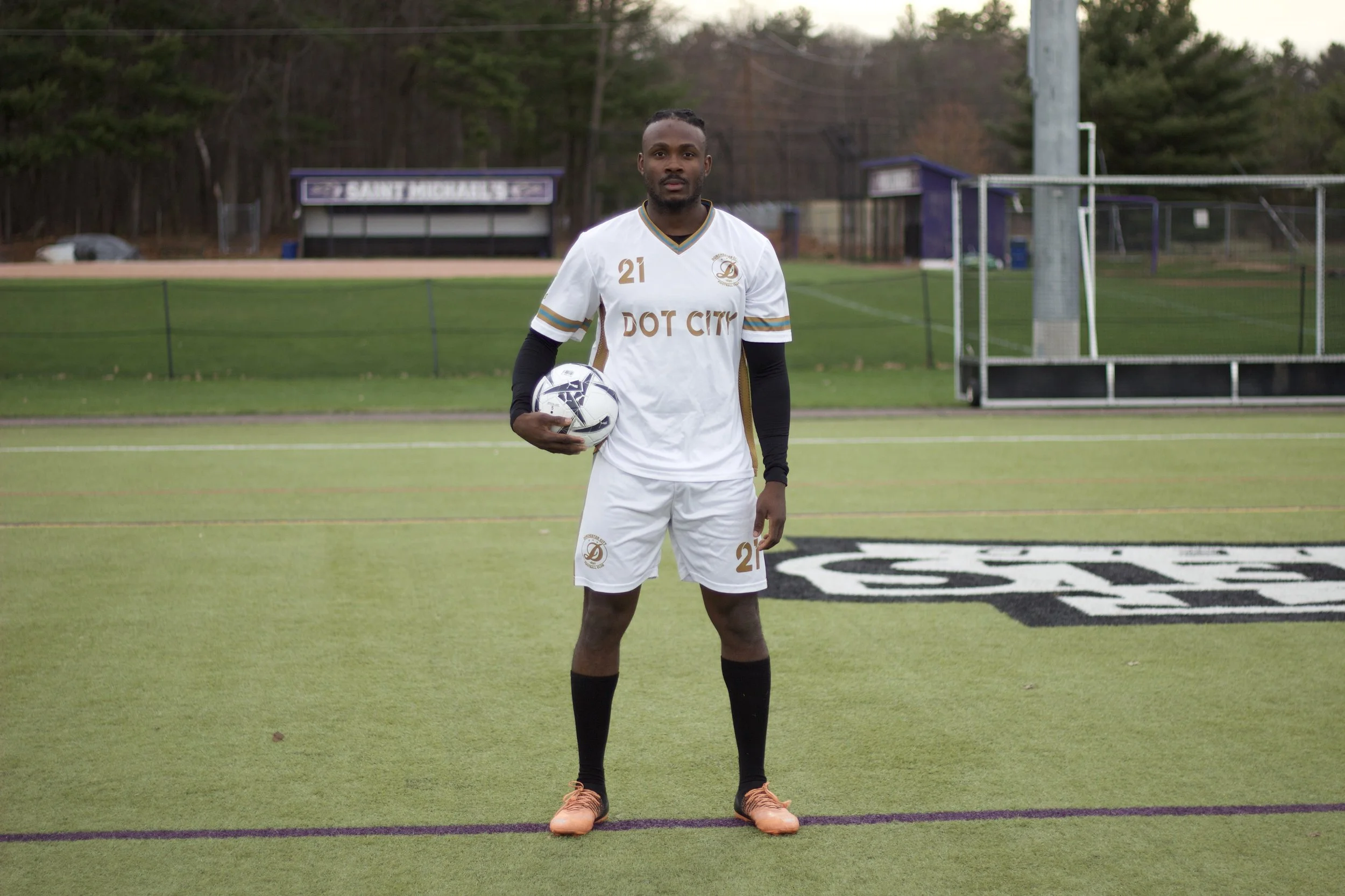 A male soccer player standing on a field holding a soccer ball, wearing a white jersey with gold accents and the number 21, with a serious expression, and an athletic build. The background includes green grass, a soccer goal, and trees.