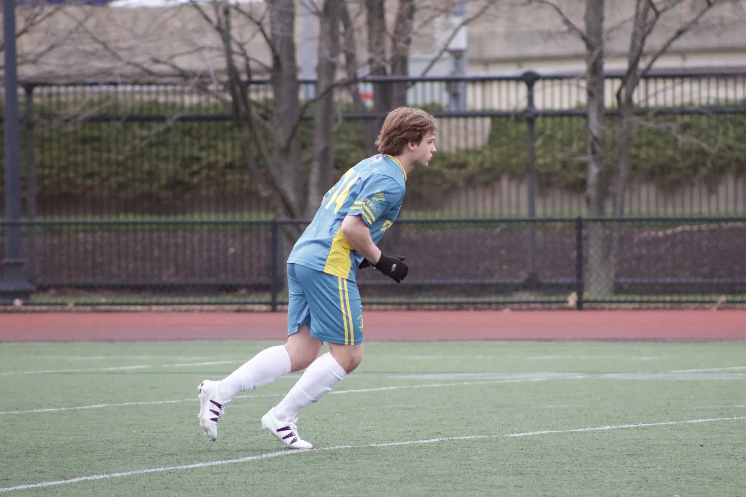 A young male soccer player with light brown hair, wearing a blue and yellow sports uniform, running on an outdoor artificial turf field during daytime. He is wearing white socks, white and purple soccer cleats, and black gloves. In the background, th