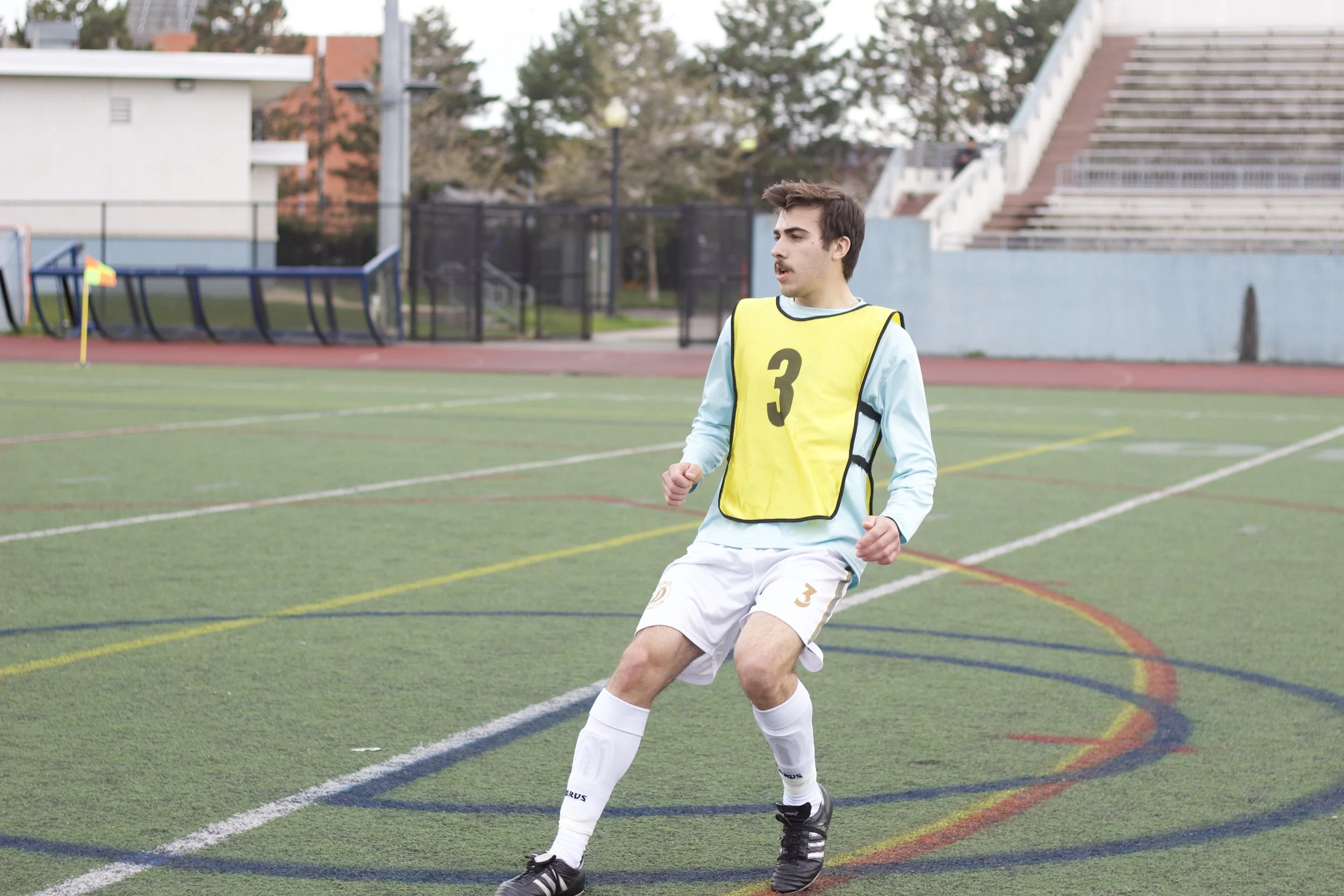A young male soccer player with dark hair wearing a yellow bib with the number 3 is on the field, mid-action, at an outdoor stadium.