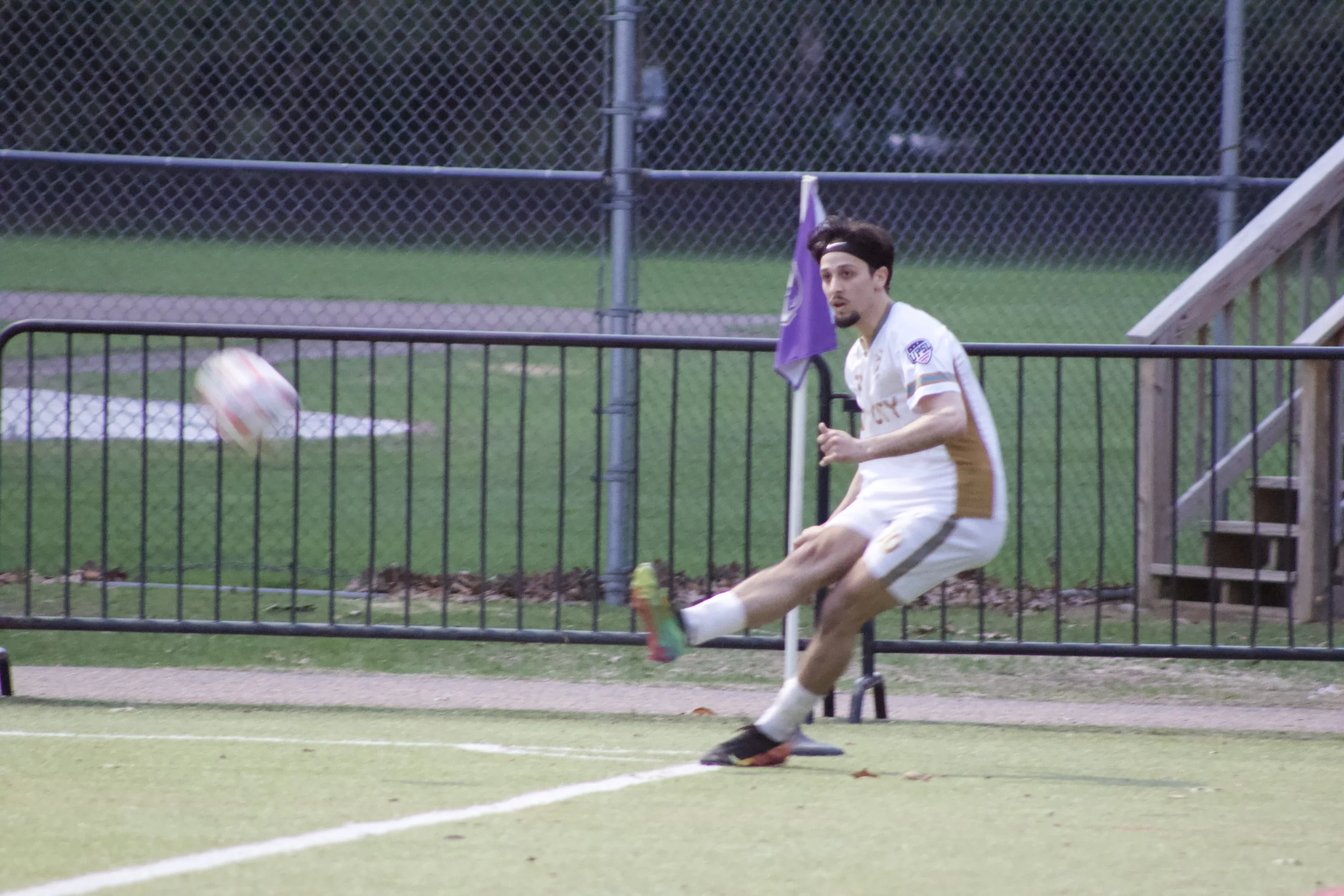 A man in a white soccer uniform kicks a soccer ball on a field near a black metal fence, with a purple flag and a set of stairs nearby.