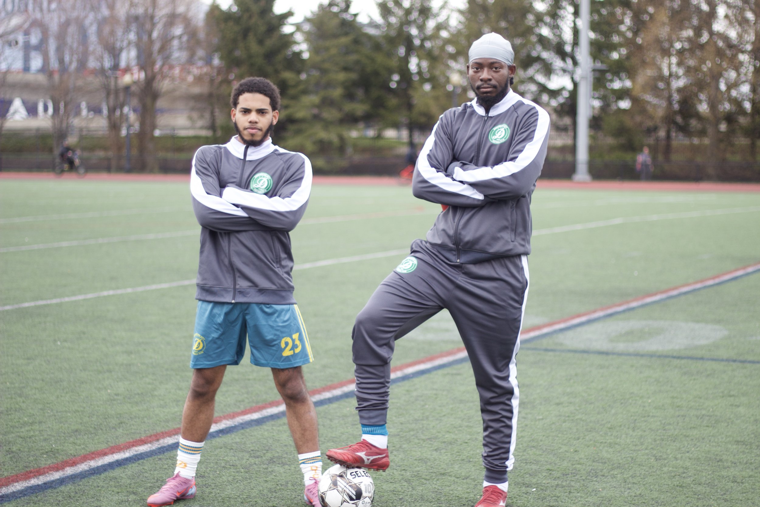 Two young men standing on a sports field with arms crossed. One is wearing a gray athletic jacket and pants, the other in a matching gray jacket and blue shorts. A soccer ball is in front of them, and they are standing on a grassy field with a runnin