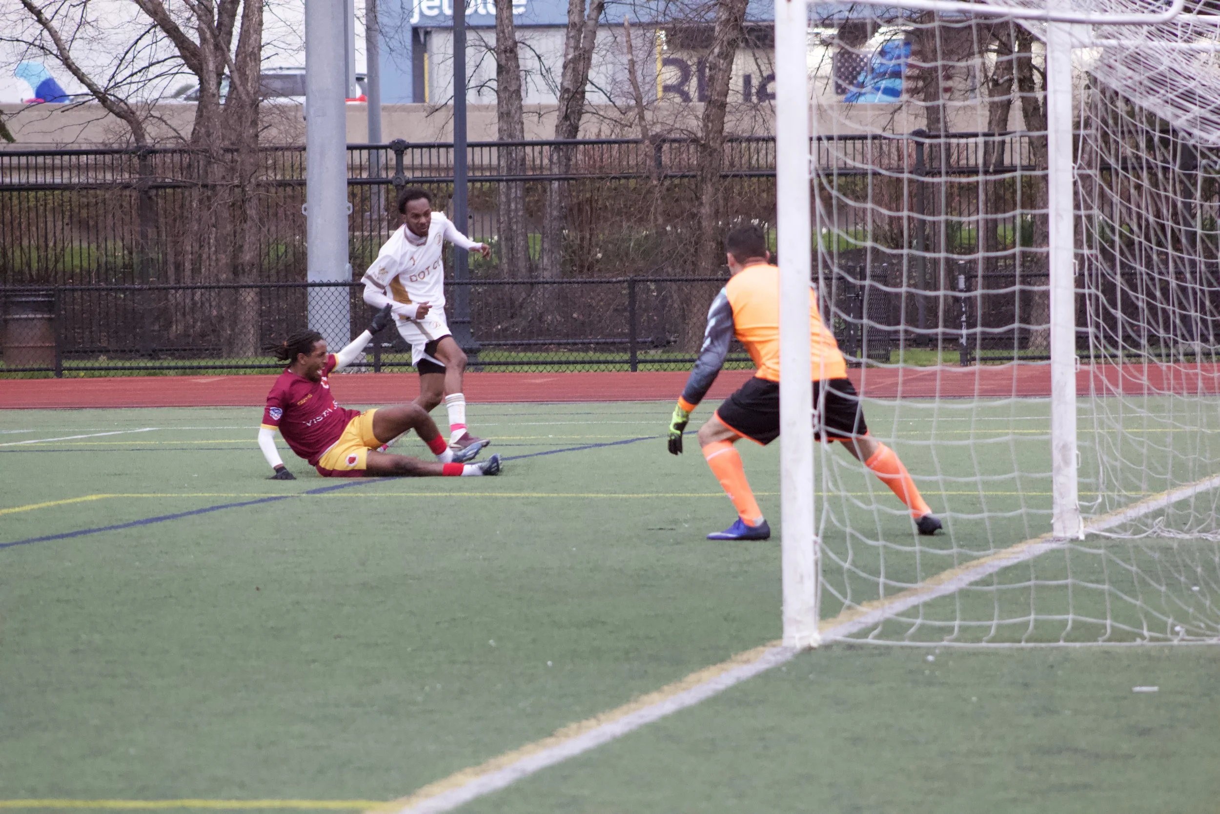 A soccer game scene with a player in a maroon uniform sitting on the ground after a collision, another player in white standing nearby, and a goalkeeper in orange ready in front of the goal. The scene is on a green artificial turf field with a fence 
