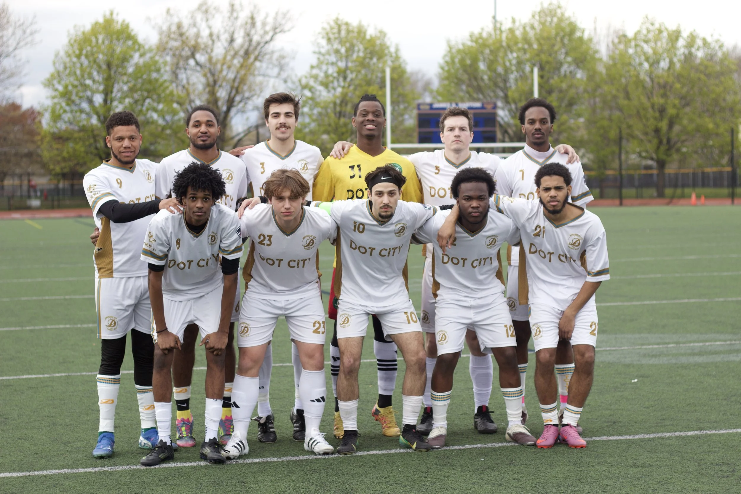 A group of young men in soccer uniforms posing together on a soccer field, with trees and goalposts in the background.