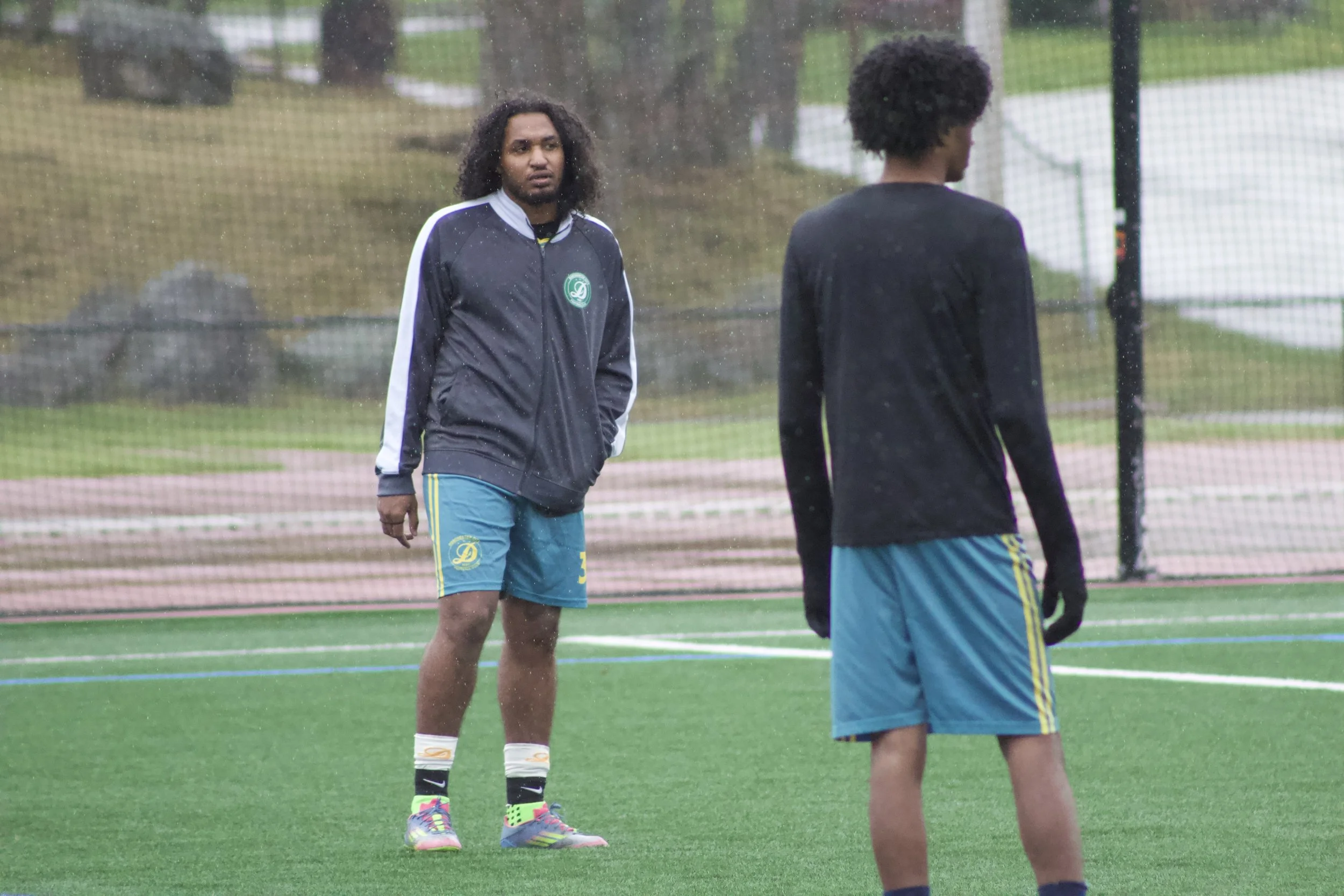 Two young men with dark curly hair standing on a rain-soaked sports field, engaging in a conversation, one facing the camera with hands at his sides and the other with his back to the camera.