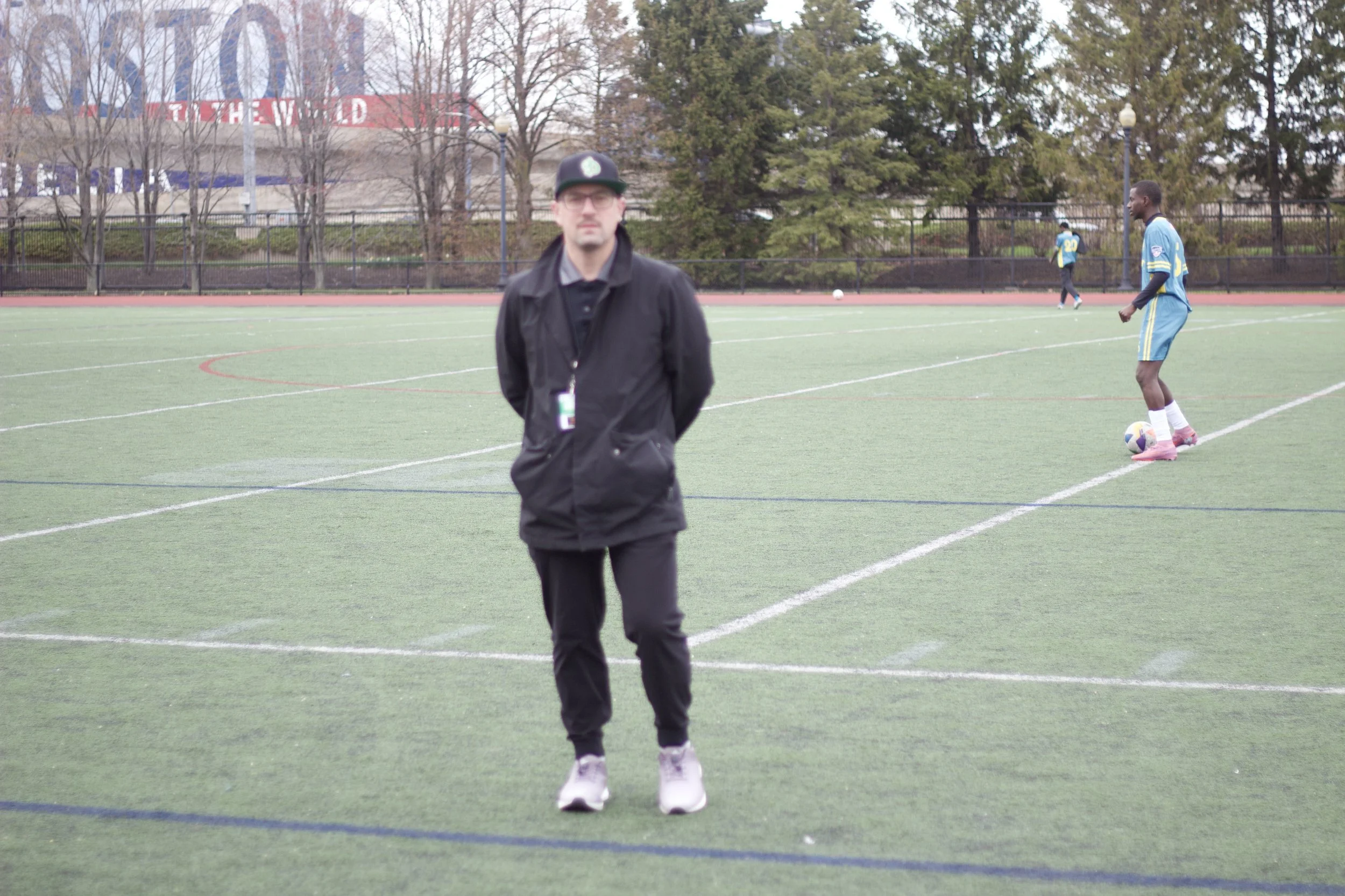 A man in a black jacket and hat stands on a football field with two players in blue jerseys and pink shoes in the background. One player is preparing to kick a soccer ball.