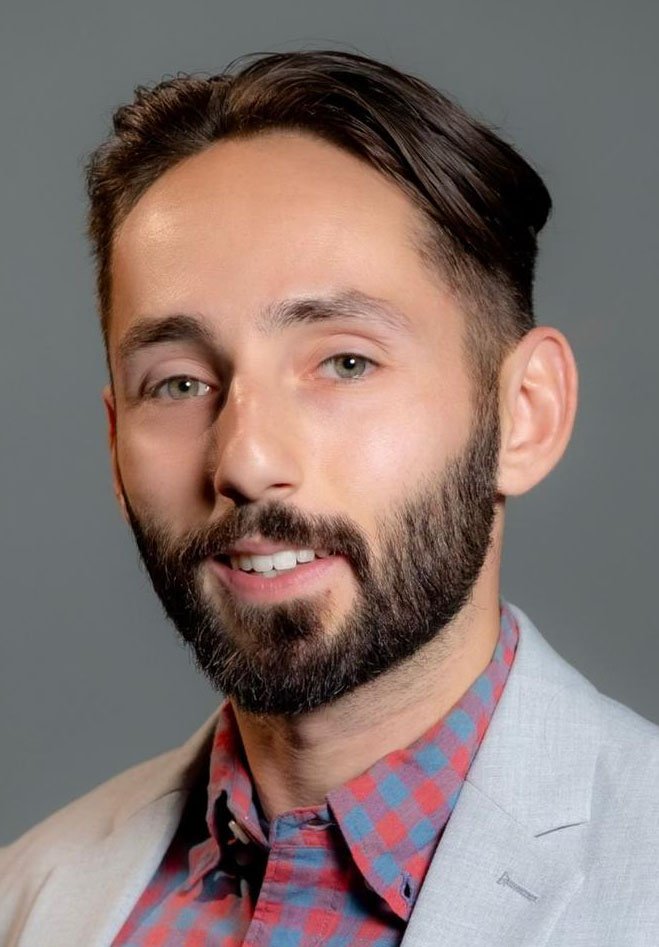 Close-up portrait of a man with dark brown hair, a well-groomed beard, light skin, wearing a light-colored blazer over a red and blue checkered shirt, against a plain gray background.