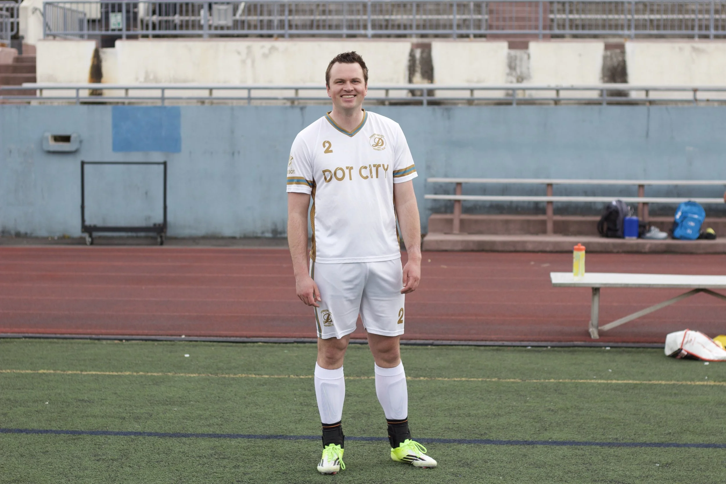 A young man standing on a soccer field wearing a white soccer uniform with gold and blue accents. He is smiling and looking at the camera, with a stadium background and equipment on the sidelines.