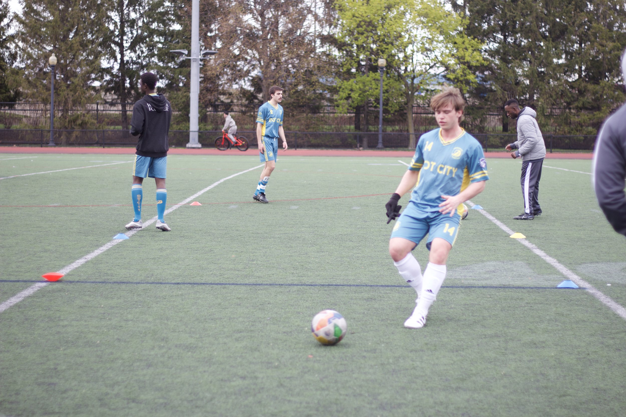 Young soccer players practicing on a field, with one kicking a multicolored ball, and others standing or walking nearby. Trees and light poles are in the background.