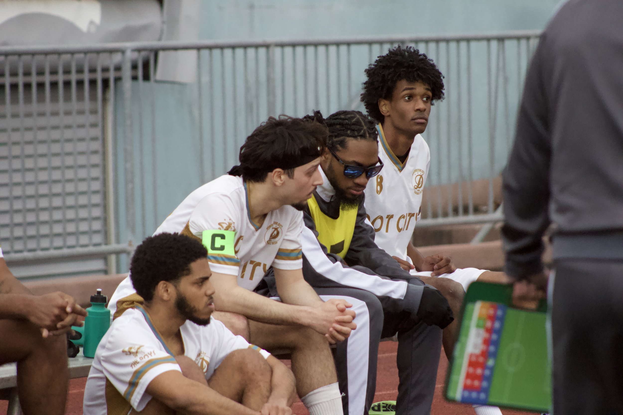 A group of soccer players sitting on a bench during a game, wearing white jerseys with gold and blue accents, and one player in sunglasses.