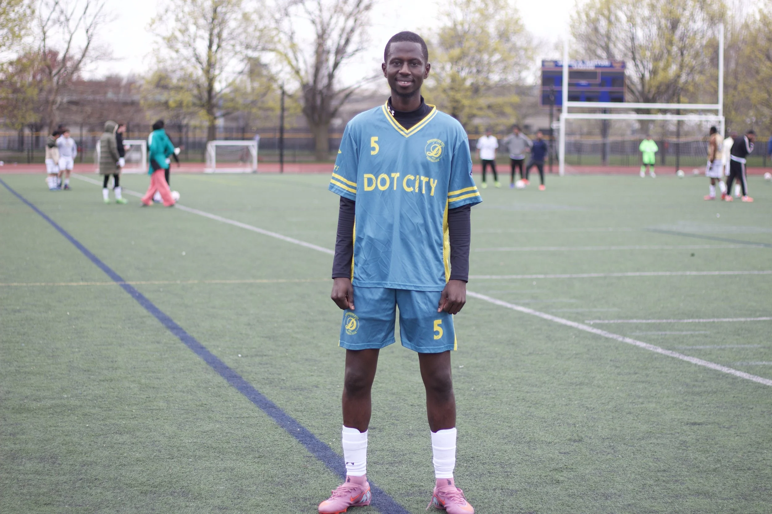 A young man in a blue soccer jersey and shorts standing on a soccer field during practice or a game, with other players and coaches in the background.