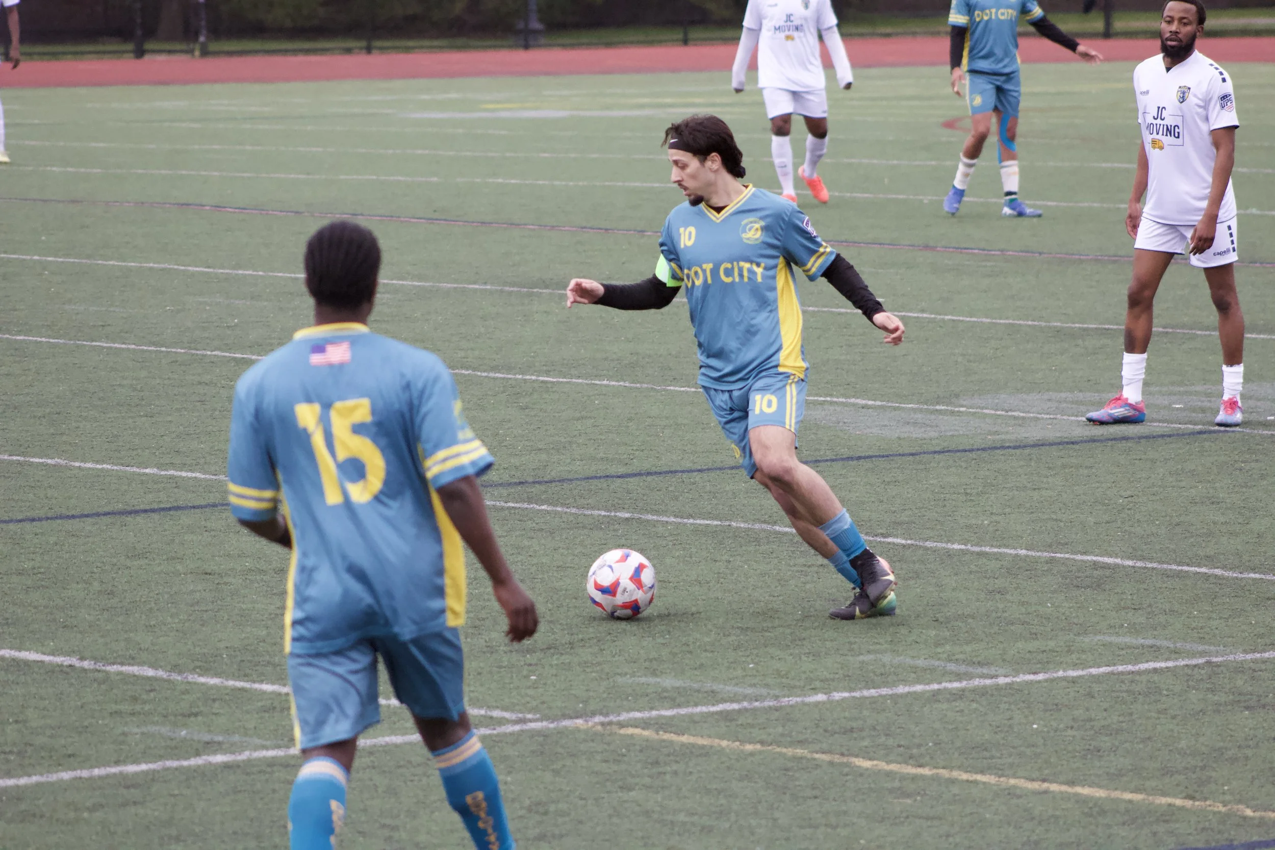 A soccer match taking place on a green field with players in blue and white jerseys. One player in a blue jersey with the number 10 is in the act of kicking a white soccer ball with red and blue patterns, while others observe. A running track surroun