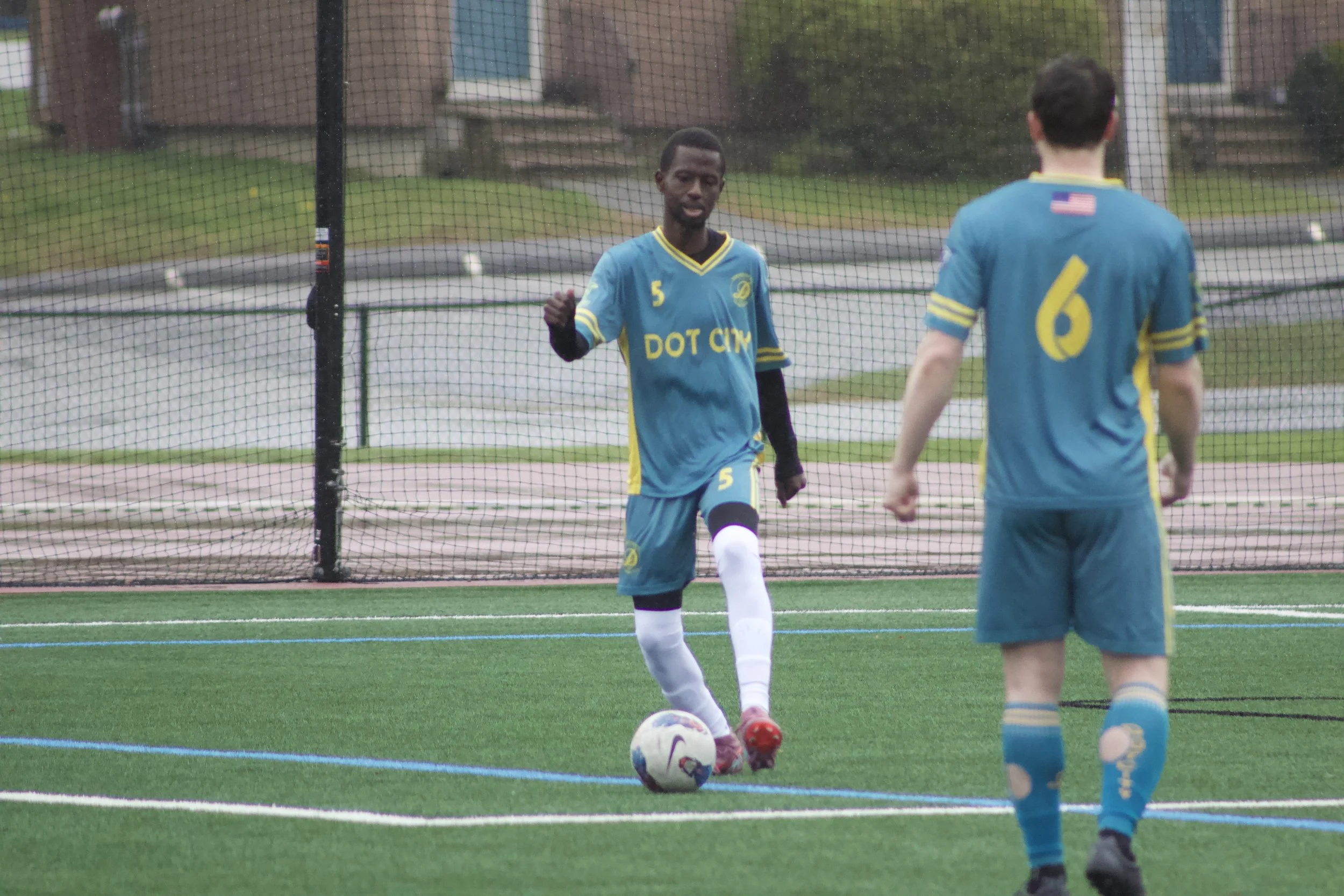 Two soccer players in blue uniforms practicing on a field near a net, with one preparing to kick a ball while the other observes, in a residential neighborhood.