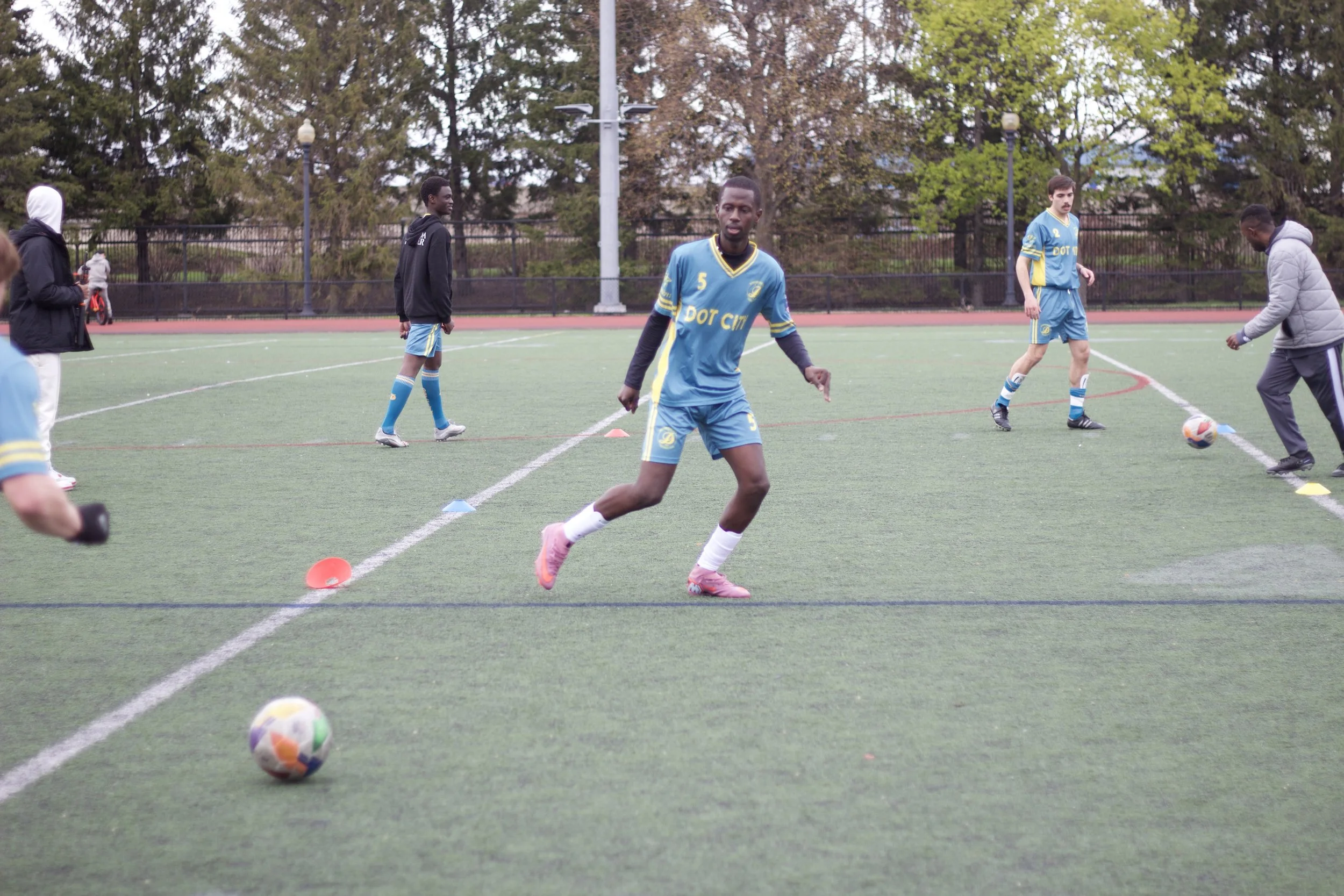 Soccer players warming up on a green field, some are stretching while a coach or trainer demonstrates with a ball.