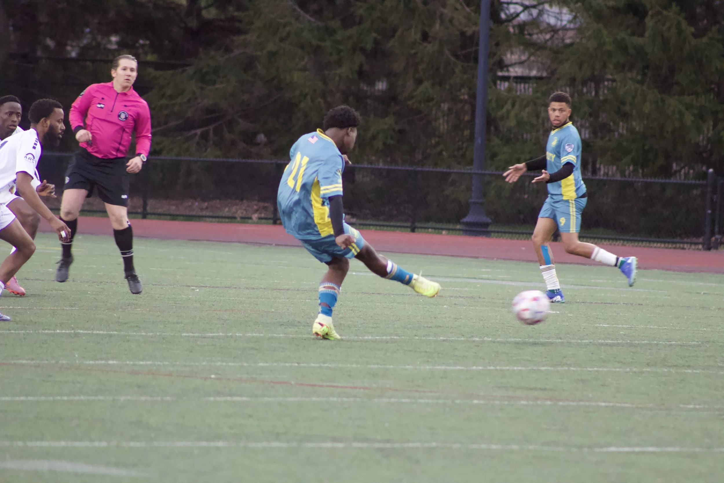 Soccer game in progress with players on the field, some wearing light blue uniforms, and an official or referee in a pink jersey.