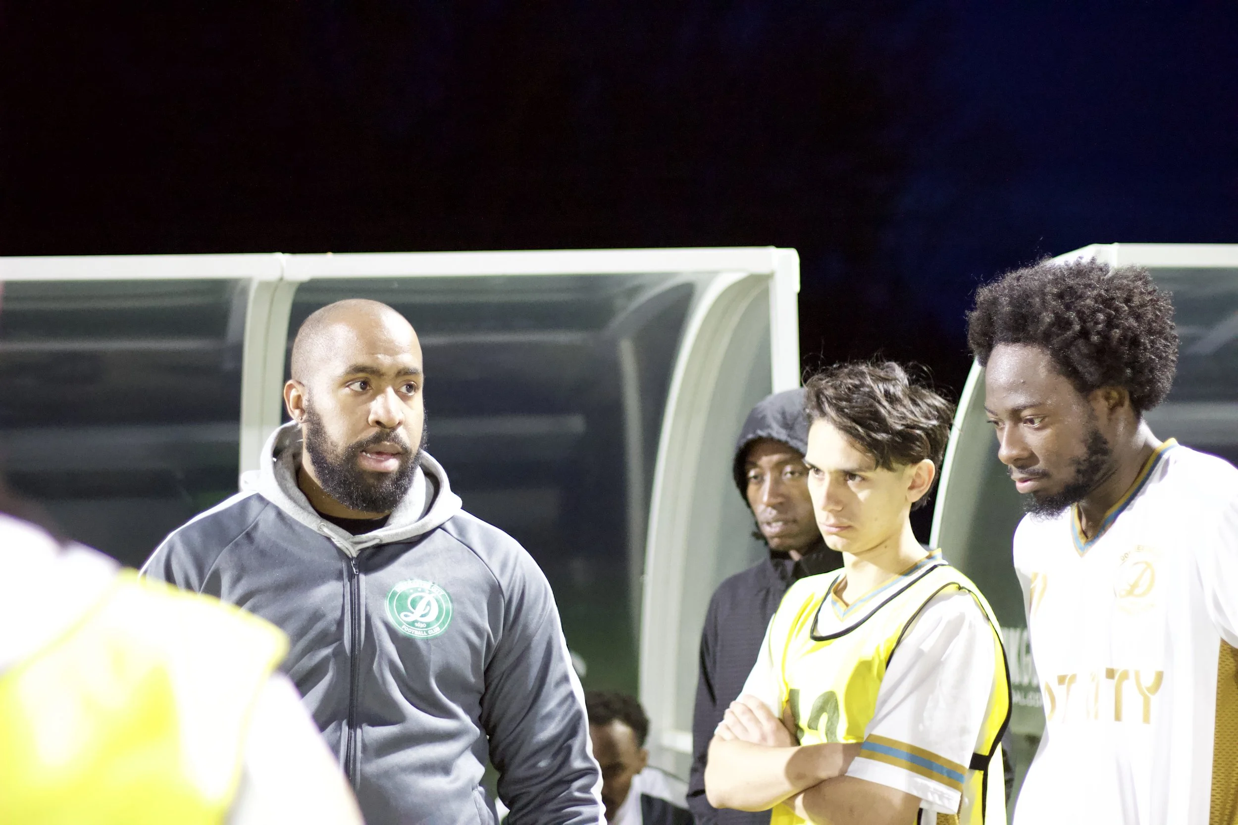 A group of soccer players and a coach standing on the sideline during a game at night, with a dark sky in the background.
