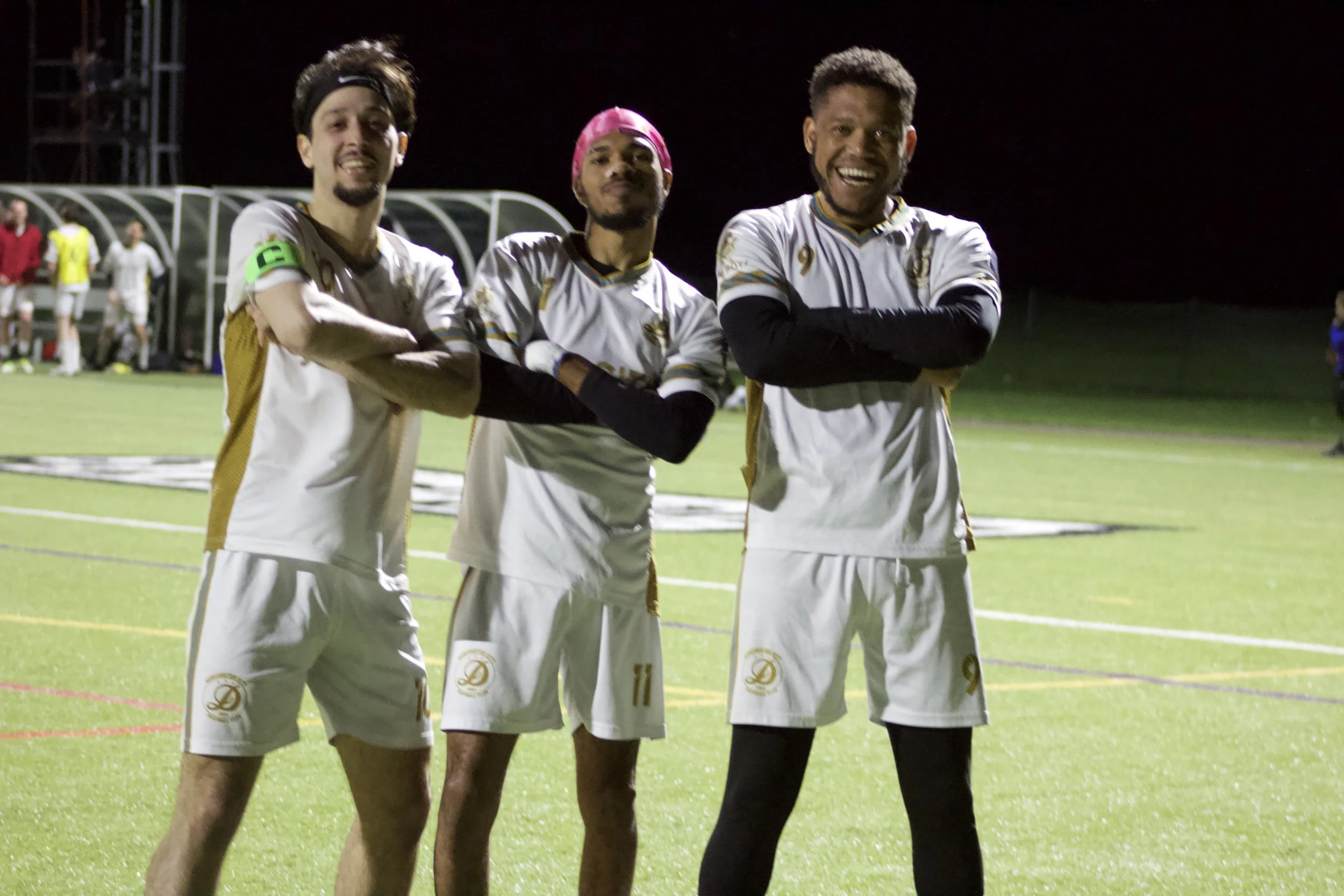 Three soccer players standing on a field during nighttime, smiling with arms crossed, wearing white uniforms with gold details.