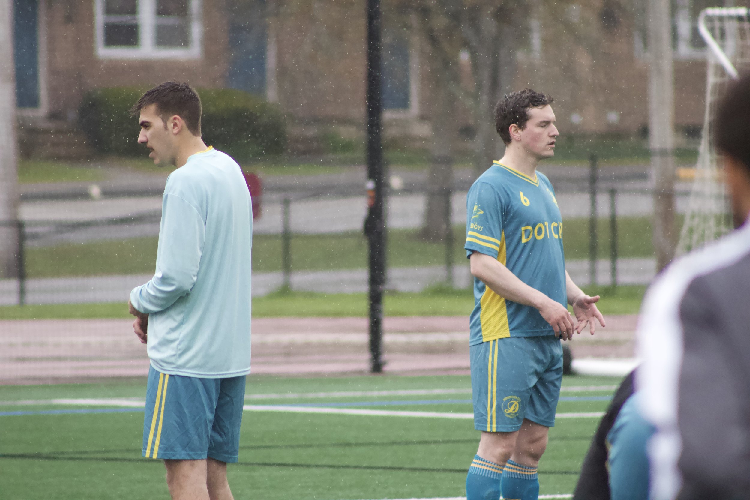 Two male soccer players standing on a field during rainy weather, facing away from each other with serious expressions.