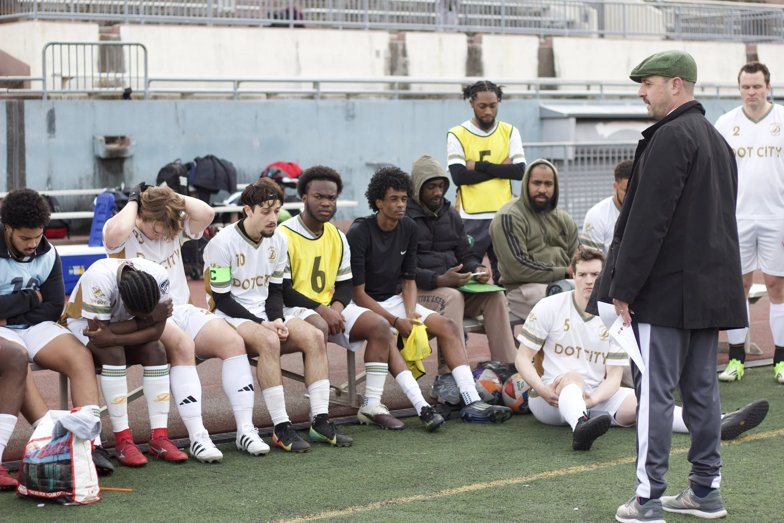 Soccer team players sitting on the bench during a game, with their coach standing in front of them.
