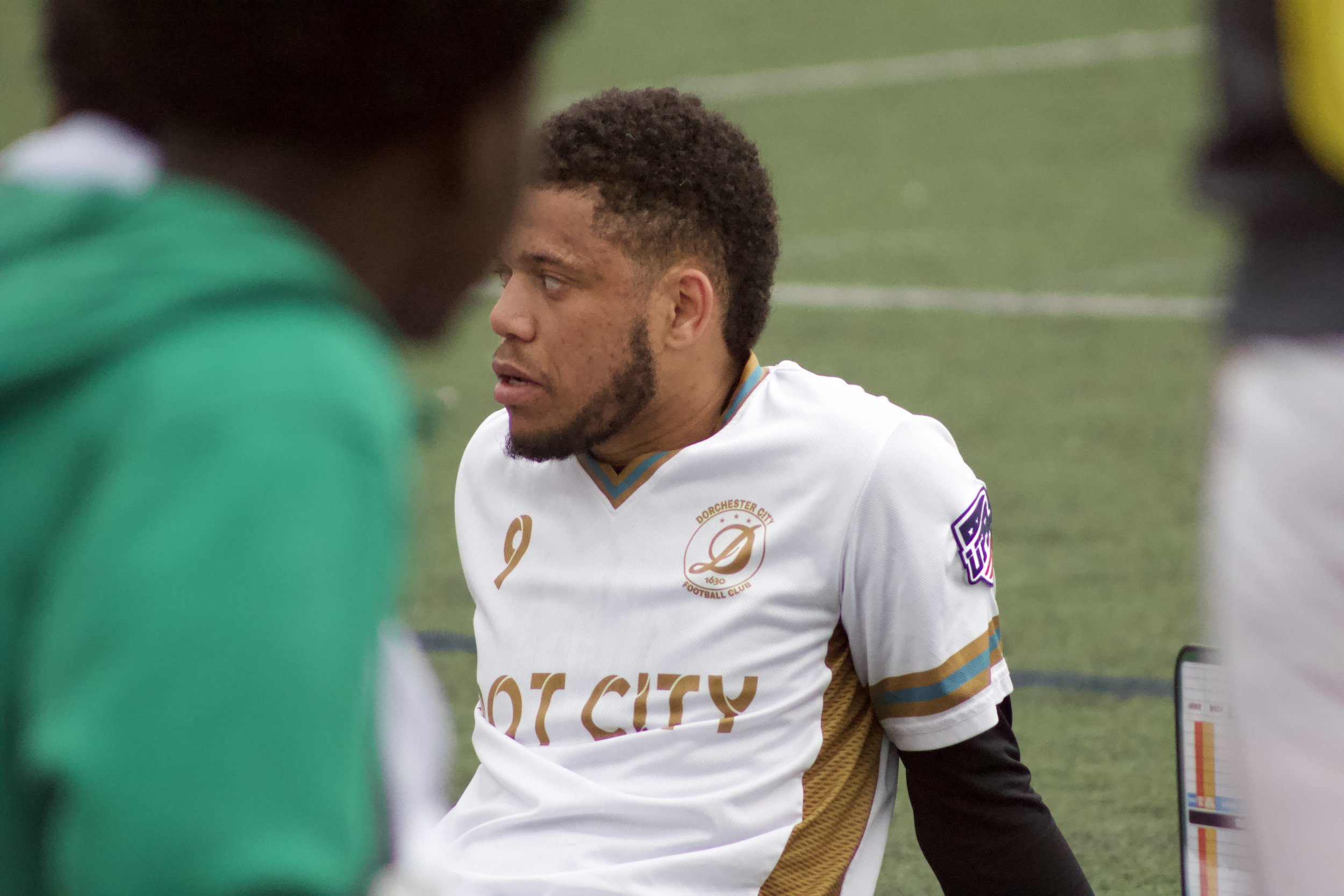 Close-up of a young man with short curly hair and facial hair, wearing a soccer jersey, looking to his left, surrounded by people on a sports field.