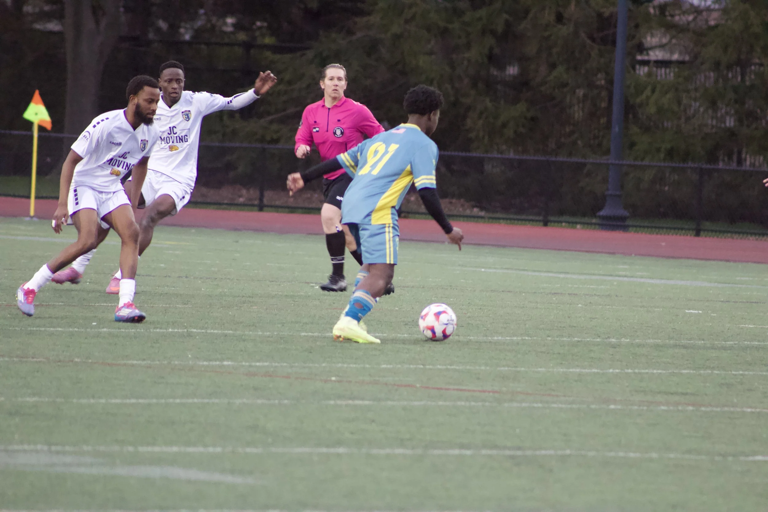 A group of soccer players competing for the ball on a field during a match, with a referee in a pink shirt observing nearby.
