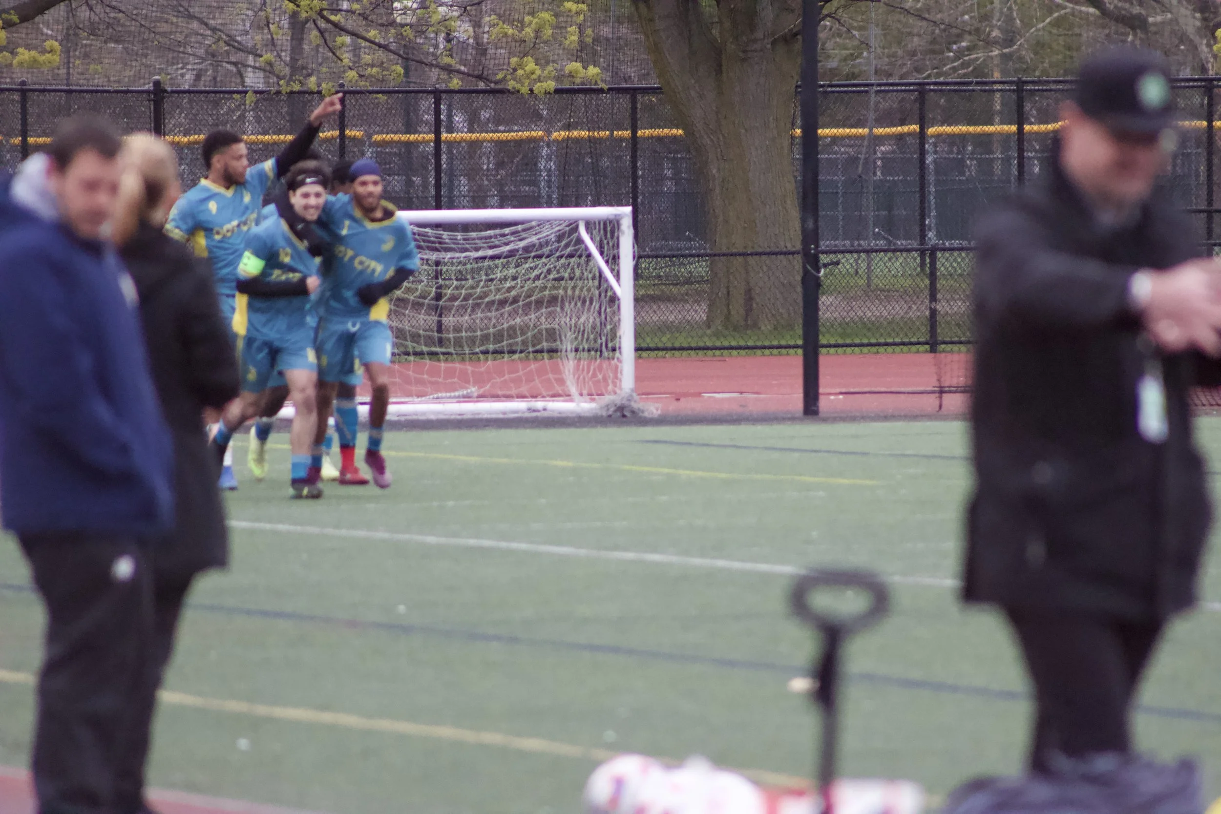A group of soccer players in blue uniforms celebrating near a goalpost on a soccer field, with spectators and trees in the background.