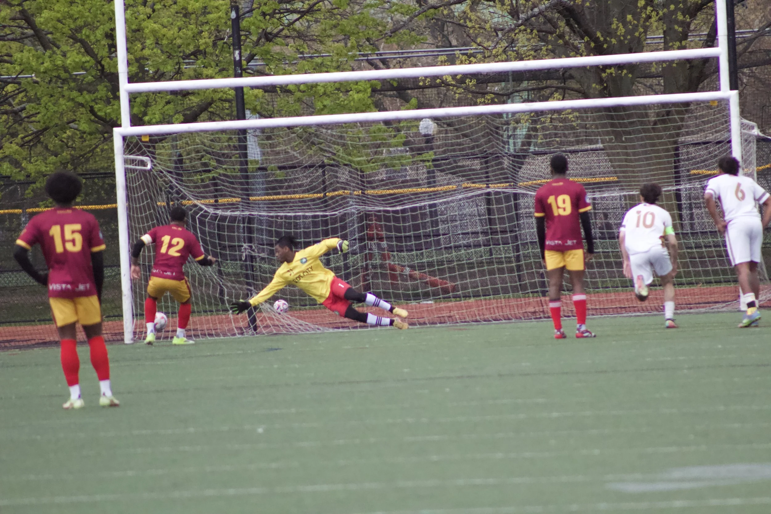 A soccer game scene showing a goalkeeper diving to save a penalty kick, with players in red and white uniforms on a field with trees in the background.
